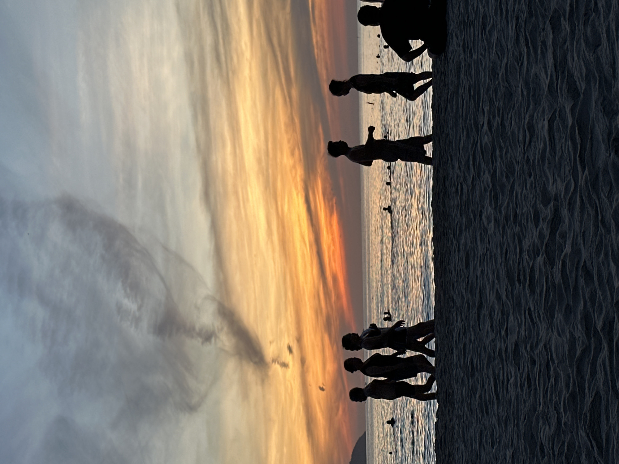 At sunset on a beach in Da Nang, Vietnam, silhouetted figures walk along the shore. The sky is painted with vibrant hues of orange, yellow, and blue, with wispy clouds forming an abstract pattern. The ocean reflects the colorful sky, creating a shimmering effect on the water's surface. Several people are visible in the water, appearing to swim or wade. The foreground is dominated by the textured sand of the beach, cast in shadow. The scene captures a peaceful evening ambiance, with the silhouettes emphasizing the natural beauty of the coastal landscape and the shared human experience of enjoying a seaside sunset.