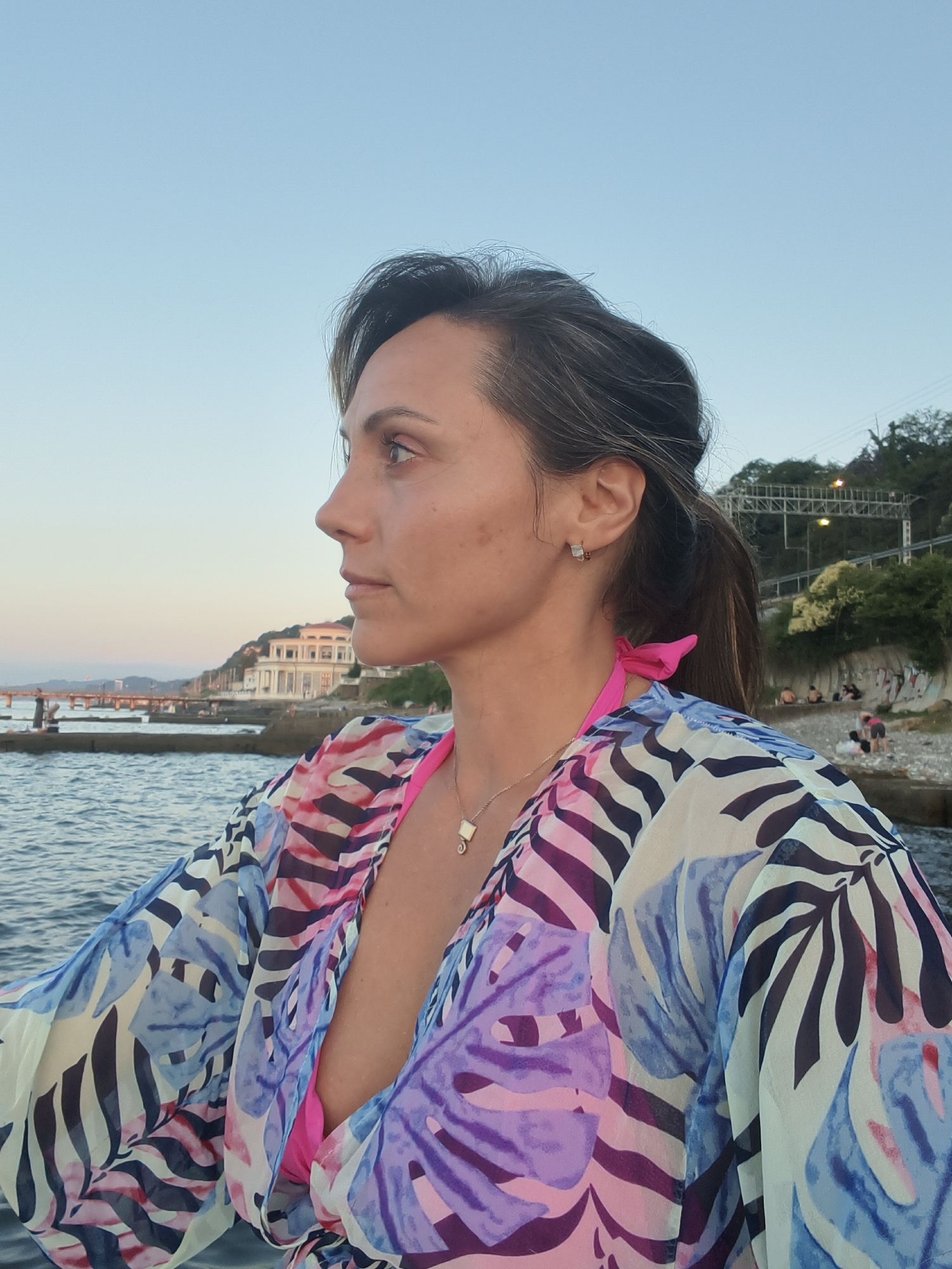 The image shows a woman in profile, facing left, with the sea and a coastal city in the background. She has dark hair pulled back in a ponytail and is wearing a colorful patterned kimono over a pink bikini top. Her expression is contemplative as she looks towards the horizon. The background features a body of water with a pier and buildings along a hillside. The overall scene appears to be a coastal resort area.
