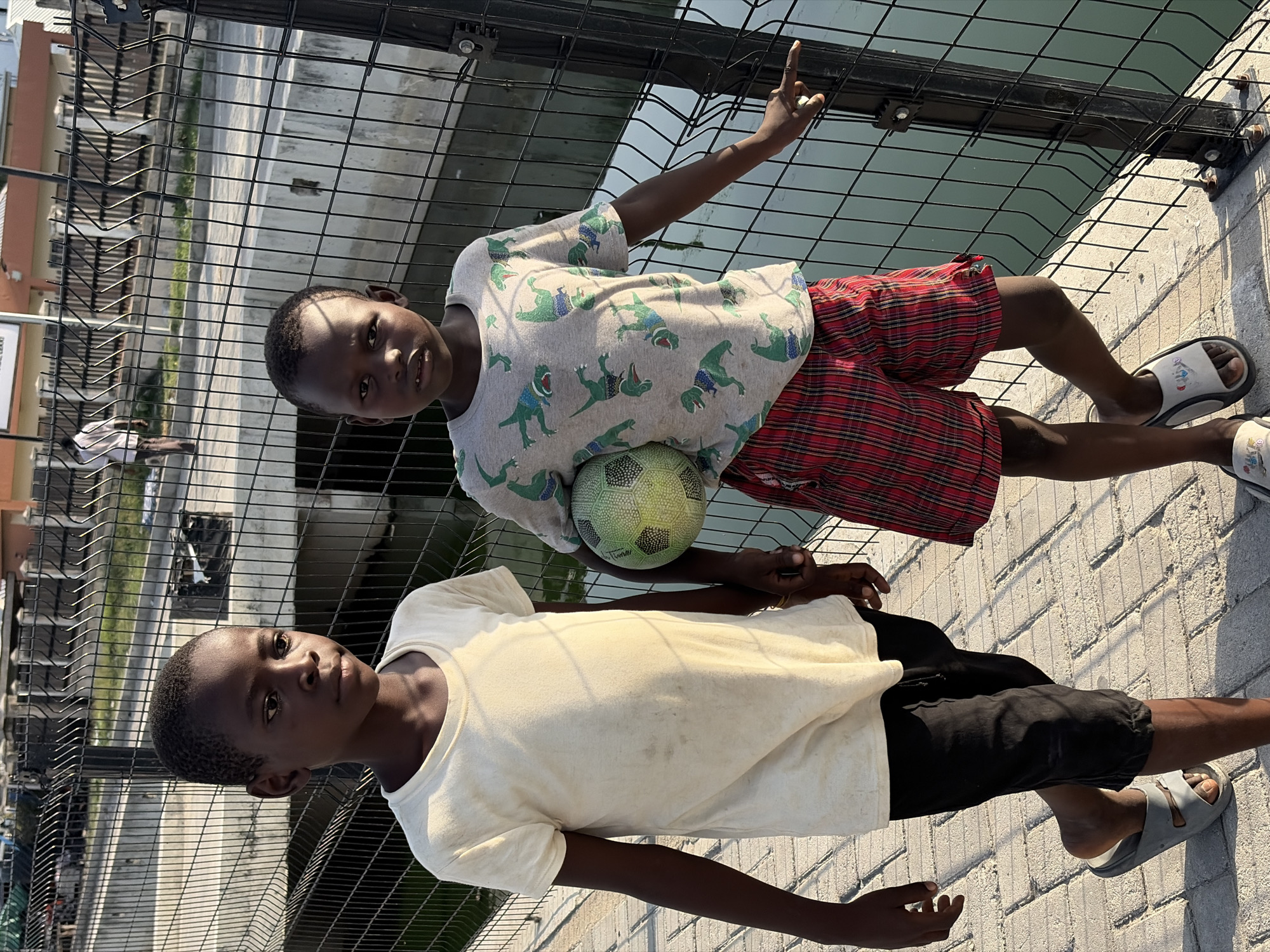 Two children are standing on a paved surface in Igboefon, Nigeria, near a fence. One child is wearing a grey dinosaur-print shirt and red plaid shorts, holding a green and black soccer ball. He points upward with his right hand. The other child wears a white shirt and black shorts. Both children are wearing sandals. A fence is visible behind them, and urban structures and vegetation are in the background.