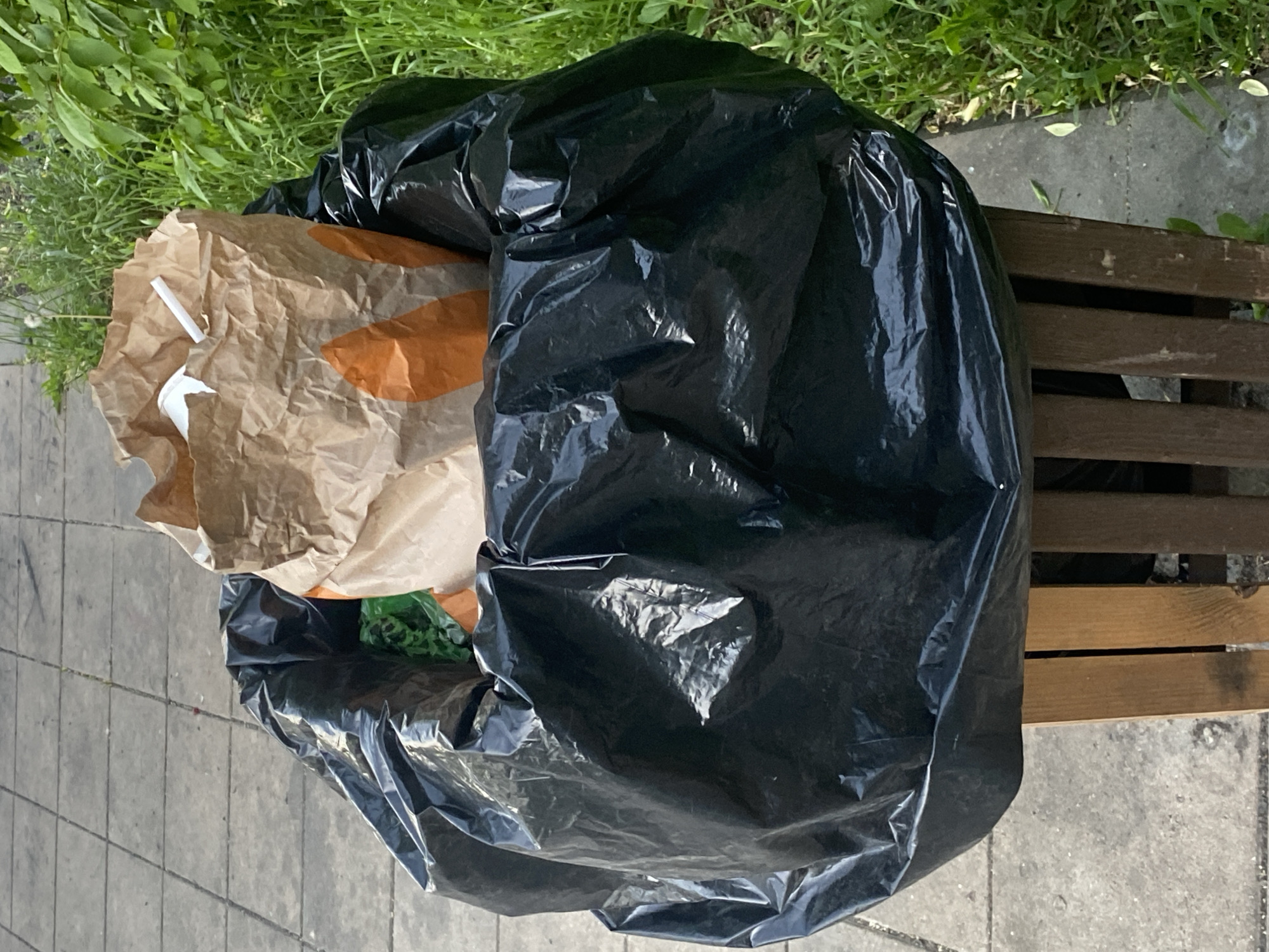 The image shows a black trash bag and a brown paper bag placed on a wooden bench. The bench is situated on a paved area, possibly a sidewalk, with grass visible in the background. The paper bag appears to contain trash, including a white plastic straw and a cup. The black trash bag is large and full, obscuring the contents. The overall scene suggests urban litter and a lack of proper waste disposal. The location in the city of Moscow, Russia, is not explicitly discernible from the image, but the scene is consistent with a typical urban environment.
