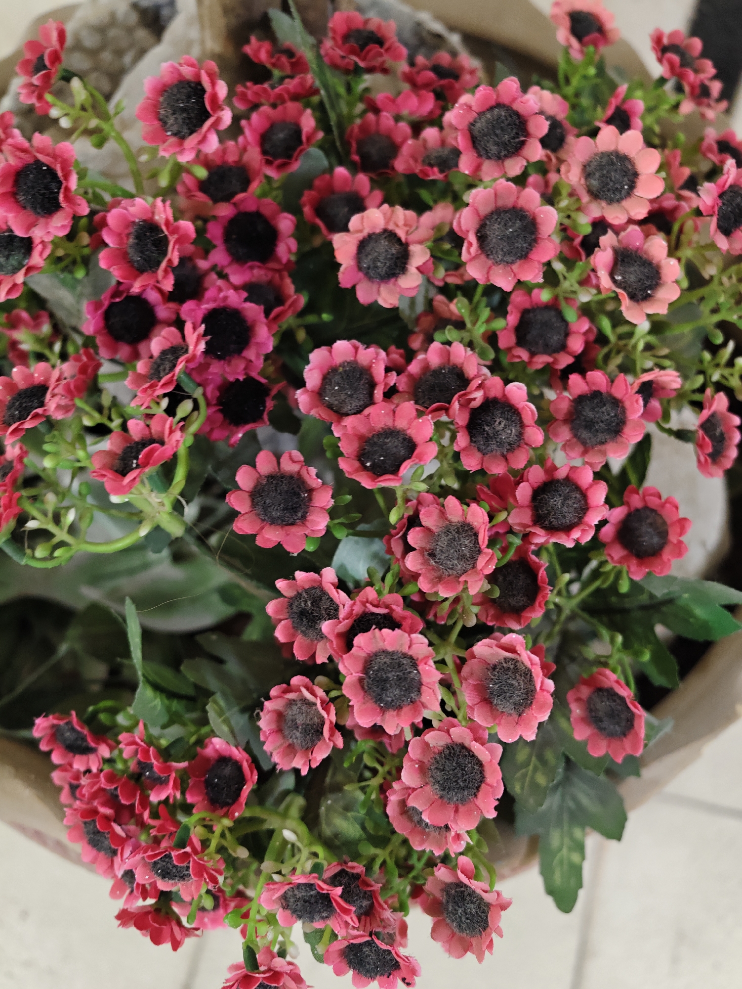 A close-up, overhead shot showcases a bouquet of small, artificial pink flowers with dark centers. The flowers are clustered together, with some facing upwards and others slightly tilted. Green artificial leaves are interspersed among the blooms. The bouquet is contained within a brown, textured material, possibly a vase or wrapping. The background is soft and out of focus, with hints of a light-colored tiled surface, suggesting an indoor setting. The image was taken in Petah Tikva, Israel.