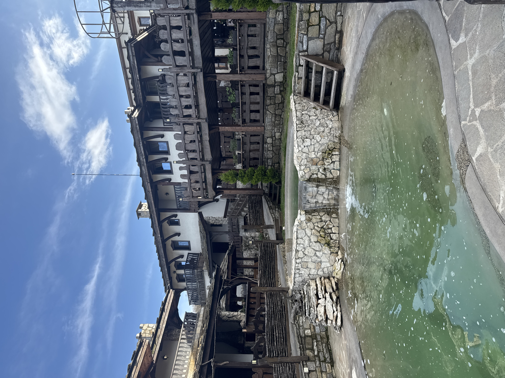 The image captures a unique architectural scene in Medovo, Bulgaria. The building is multi-storied with a distinctly rustic design, featuring extensive use of wood and stone elements in its construction, with multiple wooden balconies that add to its character. The building's design is reminiscent of traditional Bulgarian architecture.
Beside the building, there is an oval pool filled with green-tinted water. The pool's edges are lined with stonework. The sky above is a clear blue, dotted with scattered white clouds. The lighting in the image suggests it was taken during the day.