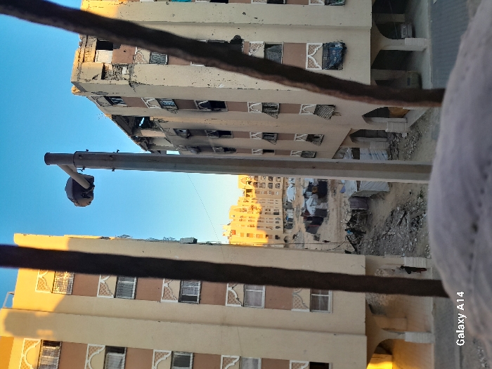 The image displays a residential street in Khan Yunis, Palestinian Territory, viewed from an elevated perspective through a blurred foreground object bearing the text "Galaxy A14" and two dark vertical bars. Two multi-story buildings with light-colored facades, brown accents, and decorative window grilles flank a central unpaved area. The building on the left appears largely intact, with its left side illuminated by direct sunlight. The building on the right exhibits significant structural damage, including compromised wall sections, shattered and fabric-covered windows, and visible debris on its upper floors.
Between these structures, the ground is unpaved, sandy, and strewn with rubble. Several makeshift shelters, constructed from tarpaulins and corrugated metal, are visible in this central area. A few individuals are discernible as small figures moving or standing near these shelters. A tall, gray street light pole with a single dark lamp head stands prominently in the mid-ground. Further back, additional residential buildings are visible under a clear, bright blue sky. A vehicle is partially seen parked beneath an archway of the right building. The scene is illuminated by direct sunlight, casting shadows that suggest either early morning or late afternoon.