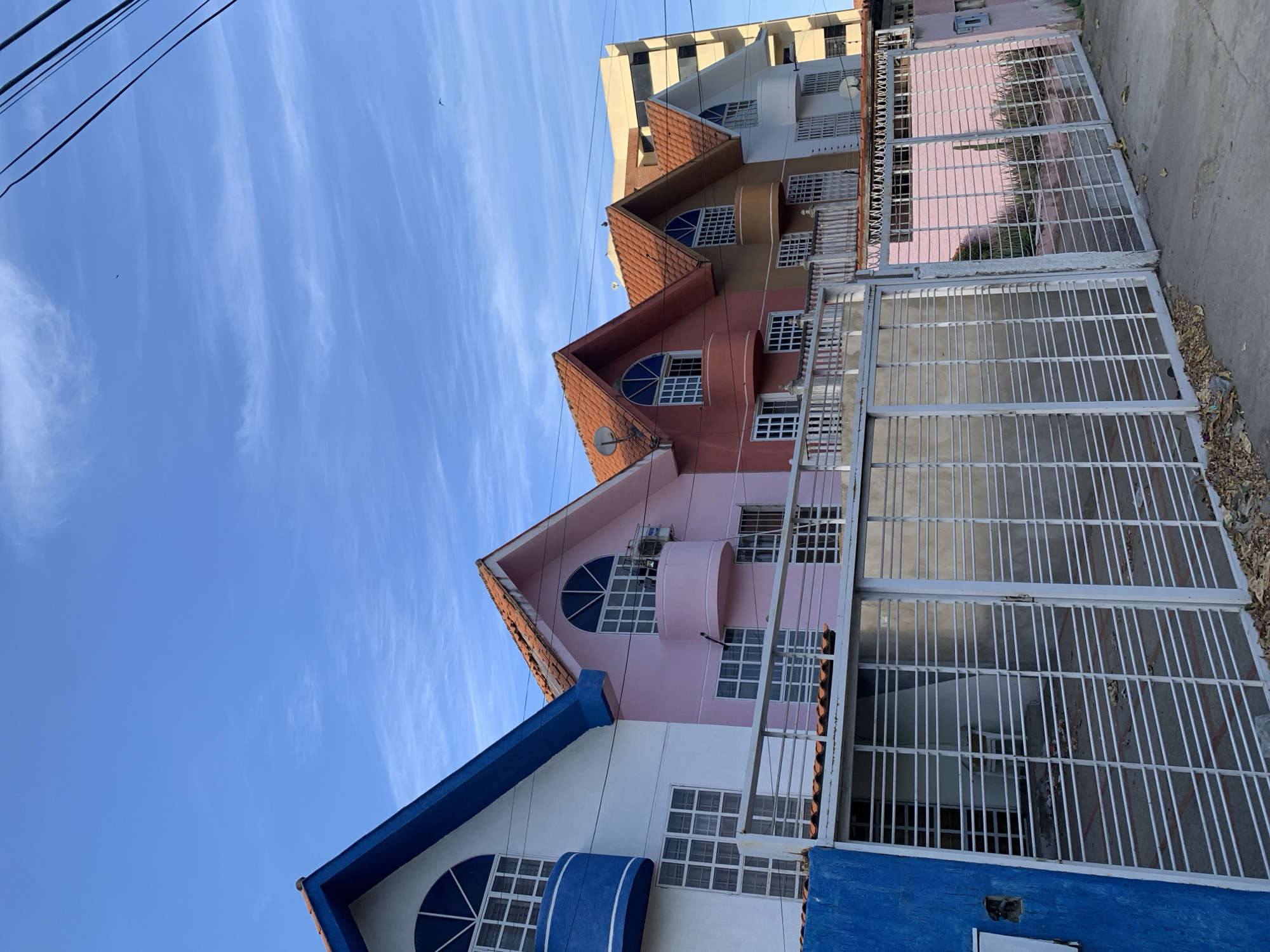 The image shows a row of colorful houses with red tiled roofs and arched windows, located in El Valle del Espíritu Santo, Venezuela. A white metal gate runs along the front of the properties. The sky is a clear blue with a few wispy clouds, and power lines are visible in the upper right corner. The houses are painted in pastel shades of white, pink, coral, and tan. A tall yellow building is visible in the background. The ground in front of the gate is paved with concrete, with some dry leaves scattered on it. The scene appears to be a residential street in a tropical or subtropical climate.