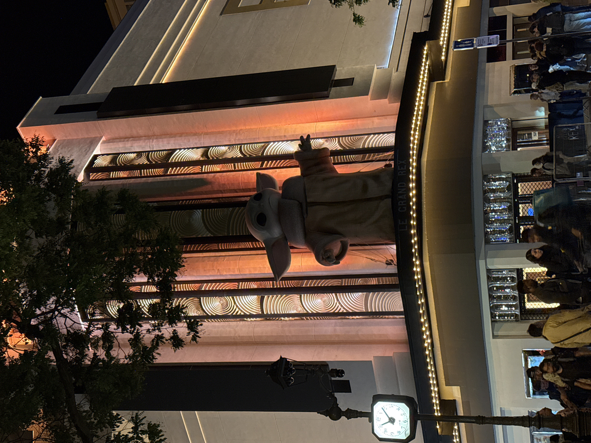 The image shows a night scene in Paris, France, featuring the facade of the Grand Rex cinema. A large "Baby Yoda" figure is displayed above the entrance. Several people are gathered near the entrance of the building. A street clock is visible in the foreground. The cinema's architecture includes decorative circular patterns and linear lighting.