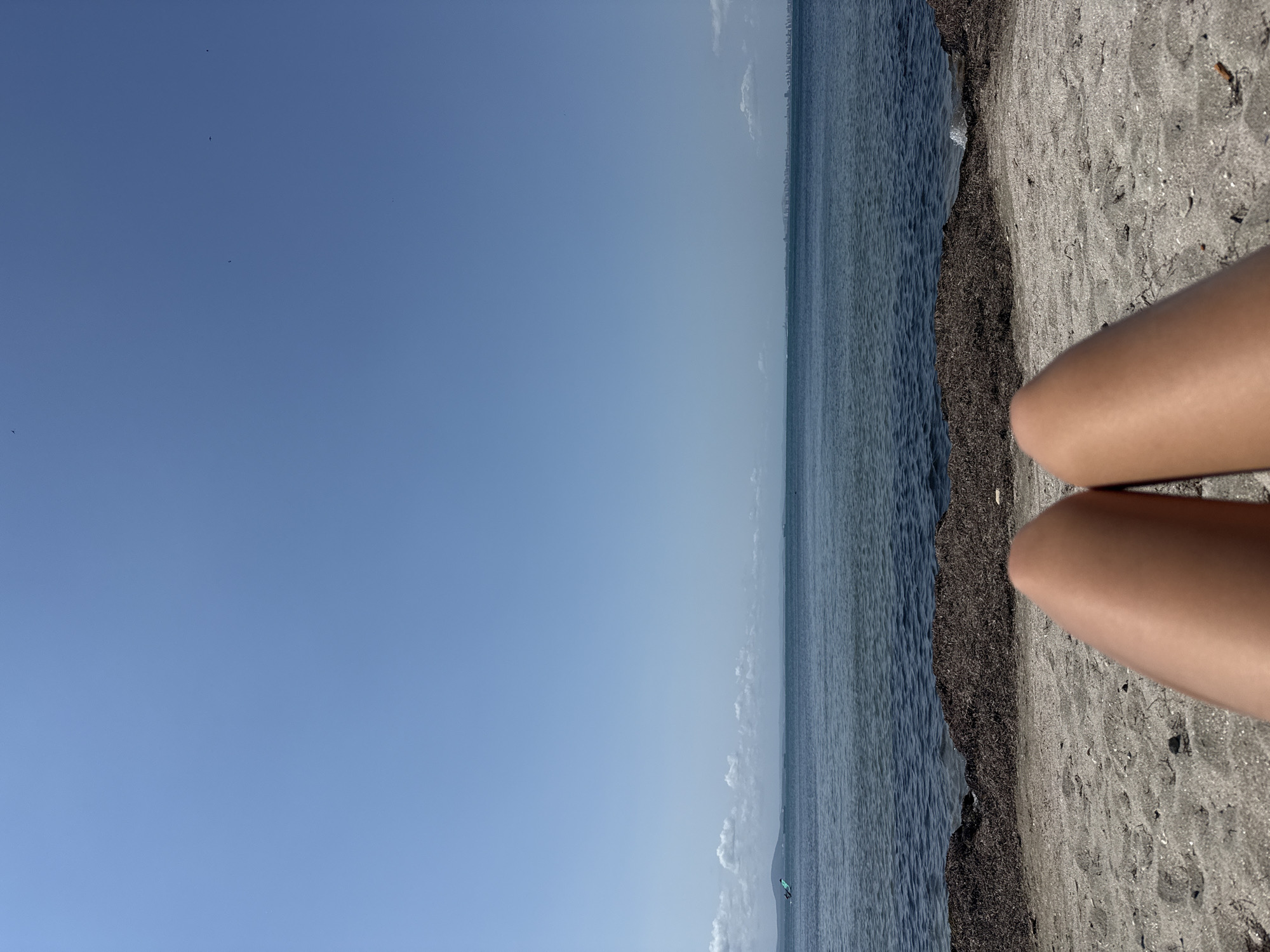 The image depicts a serene view of the sea from a sandy beach in Pomorie, Bulgaria. The foreground is dominated by the sun-kissed legs of a person, positioned as if relaxing on the shore. Beyond the sandy expanse, a dark line of seaweed marks the transition to the calm, deep blue water. In the distance, a hazy horizon reveals the outline of land, possibly a coastline, and hints of distant structures, suggesting a populated area. The sky above is a vast expanse of clear, bright blue, interrupted only by a few wisps of white clouds near the horizon. The overall impression is one of peaceful solitude and enjoyment of a summer day by the sea.