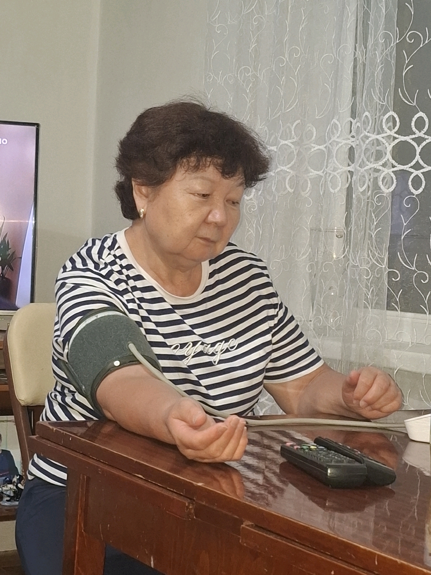 An elderly woman with short, dark hair is seated at a wooden table, taking her blood pressure. She is wearing a striped t-shirt and dark pants, and her left arm is fitted with a blood pressure cuff. Two remote controls are visible on the table in front of her. In the background, a television screen is partially visible to the left, and a sheer curtain with an embroidered floral pattern is on the right. The scene appears to be indoors, likely in a home setting in Almaty, Kazakhstan.