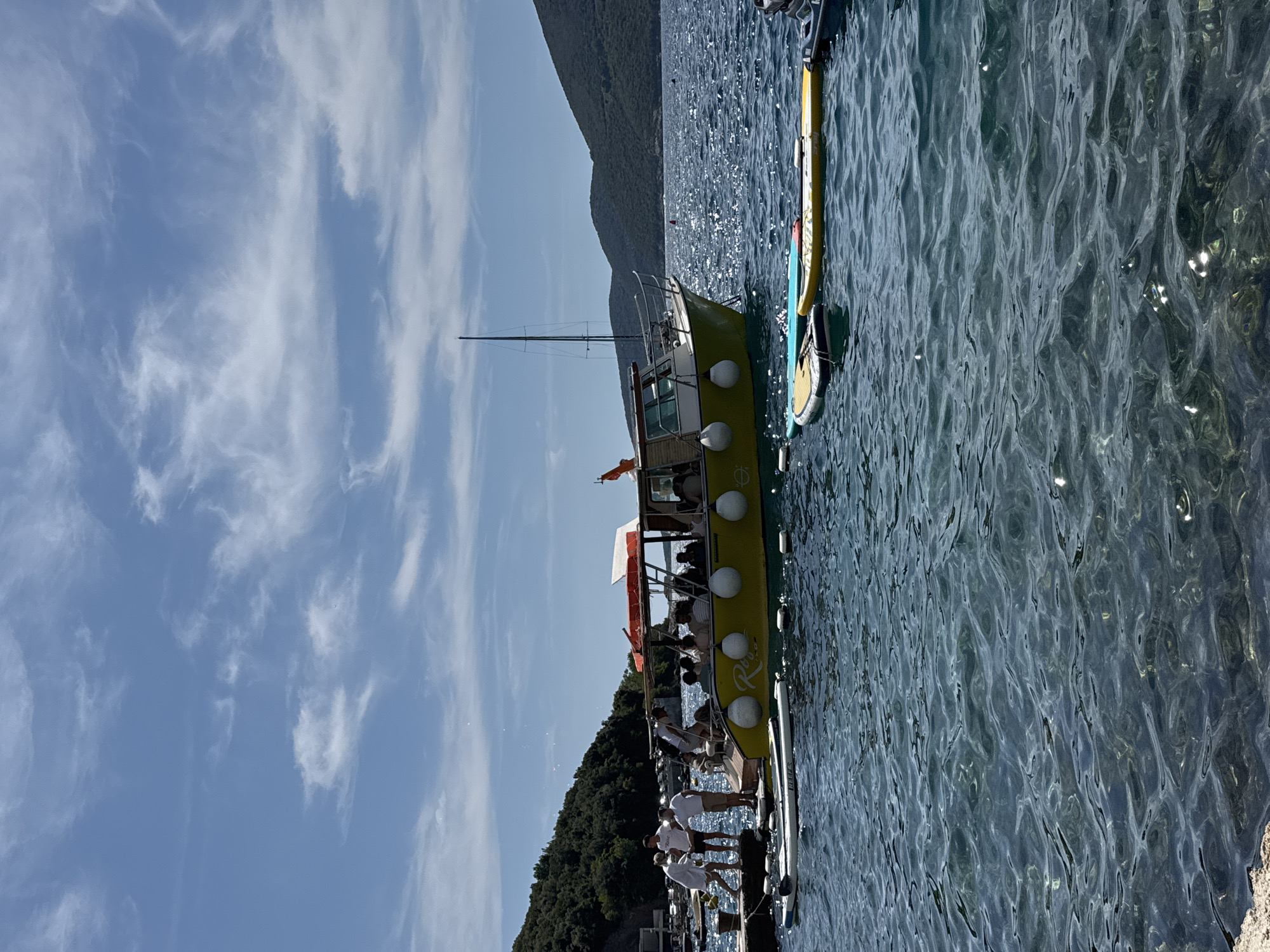 The image depicts a bright, sunny day at a harbor. A yellow passenger boat, with people visible on deck, is docked next to a pier. Several stand-up paddleboards are floating in the clear, blue water. In the background, lush green hills rise from the water's edge, and the sky is a vibrant blue with scattered white clouds. The scene suggests a recreational setting, possibly a tourist area or a beach resort.