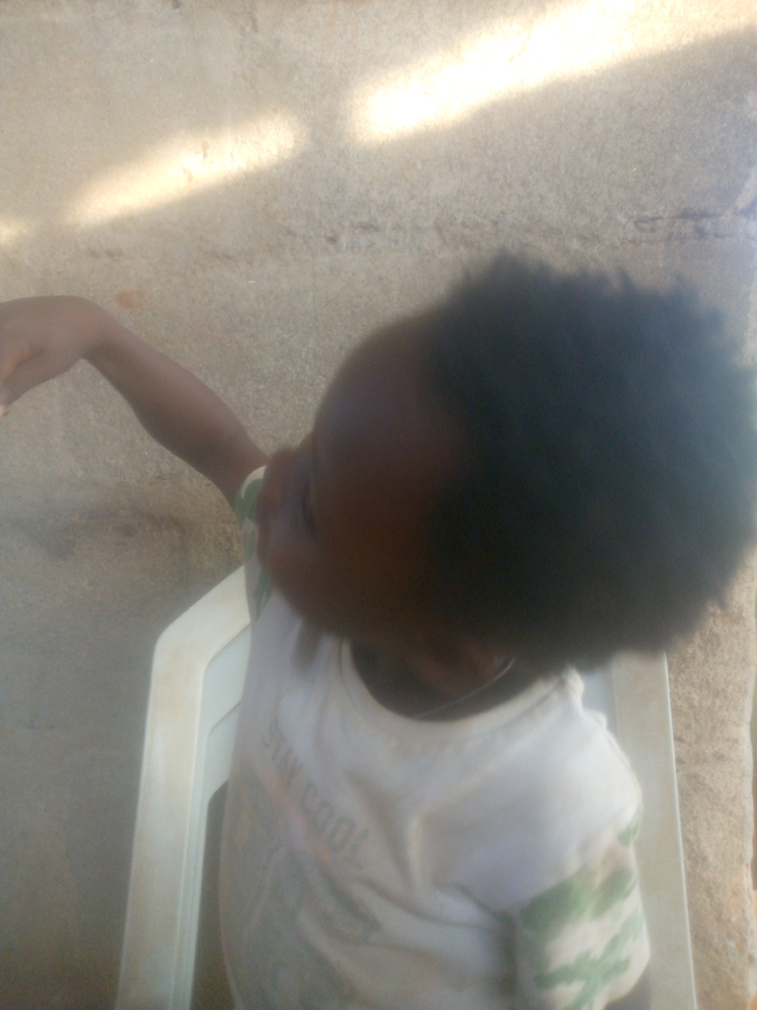 A child with dark hair sits in a white plastic chair. The child wears a white t-shirt with green and orange markings, and their right arm is raised. The location appears to be outdoors, with a light-colored, rough surface visible in the background. The setting is in the city of Unknown, Unknown.