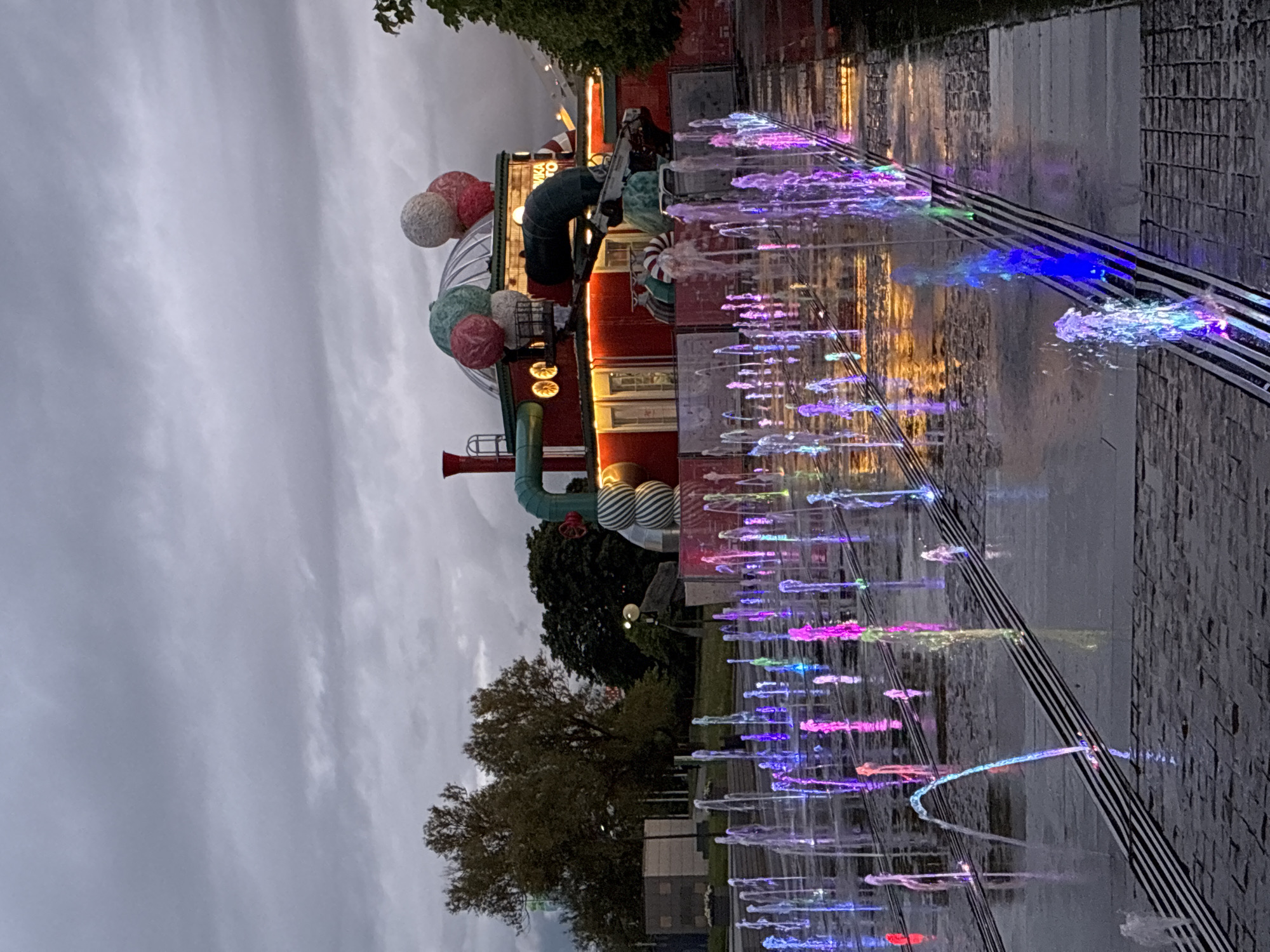 The image shows a view of the Central Children's Store on Lubyanka Square in Moscow, Russia, at dusk or in the early evening. The building is illuminated with colorful lights and festive decorations, including large decorative spheres resembling balloons attached to the building's façade. In front of the building is a fountain with illuminated water jets, creating a colorful display. The ground is wet, likely from the fountain or recent rain. The sky is overcast and grey. A few trees are visible in the background. There are no people visible in the immediate foreground.
