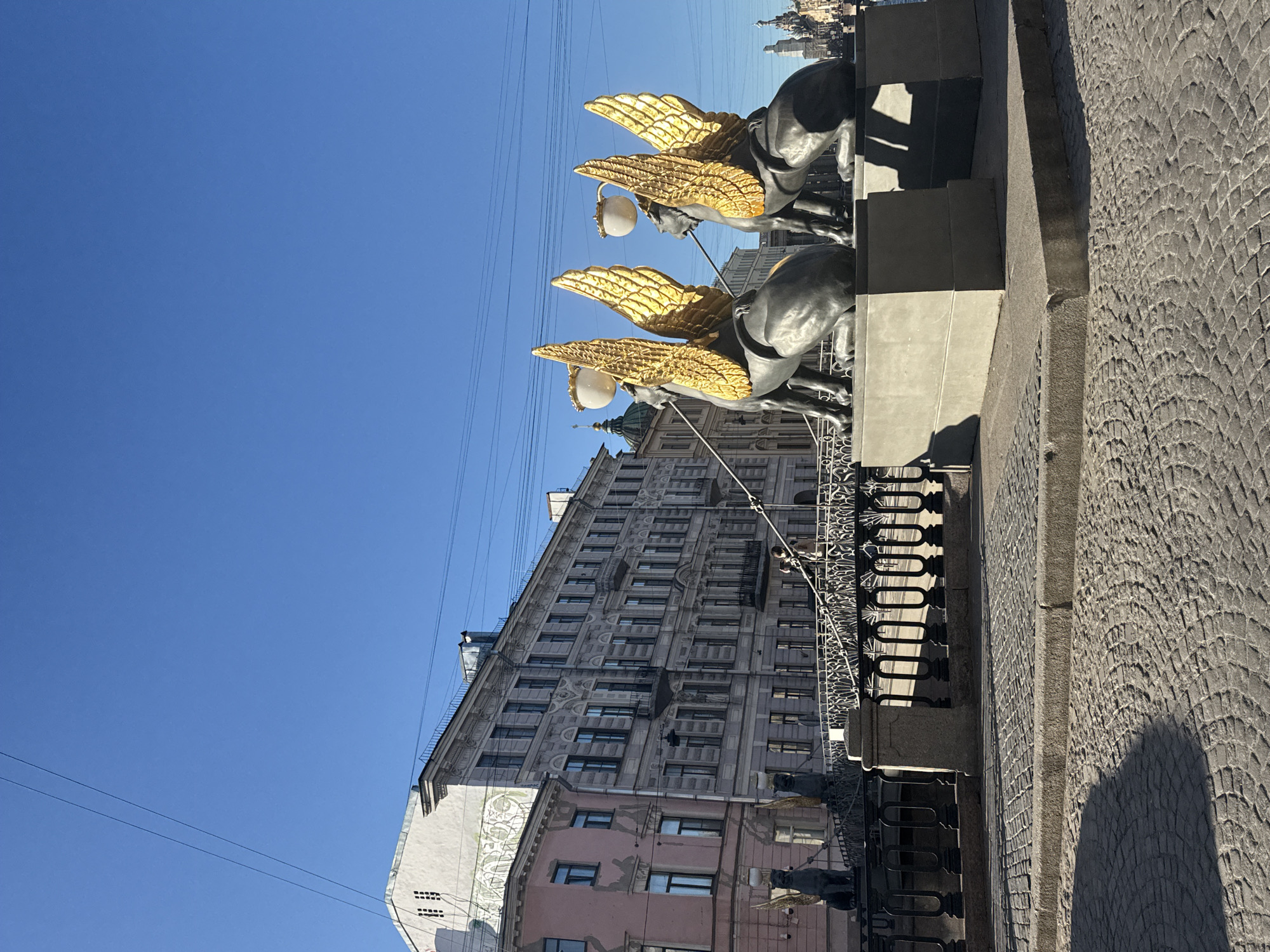 The image, taken on a clear, sunny day, depicts a section of a bridge adorned with four majestic statues of winged lions, also known as griffins. These statues, facing towards the viewer's left, feature prominent golden wings contrasting with their dark, metallic bodies. Each griffin holds a spherical, white lamp. A dark metal railing runs behind the statues, forming part of the bridge's structure.
To the right, a textured cobblestone path or roadway is visible, marked by a deep, curved shadow cast by the bridge or an unseen structure, indicating strong, direct sunlight. The sky is a vibrant, cloudless blue, traversed by several thin utility wires.
In the background, a tall, multi-storied building with intricate architectural details and numerous windows stands prominently. Below it, parts of other buildings are visible, including one with a lighter, decorative facade and another with a soft pink hue. The scene is static, with no discernible activity or people. The overall impression is one of historical grandeur in an urban environment, characteristic of Saint Petersburg, Russia. There is no visible text in the image.