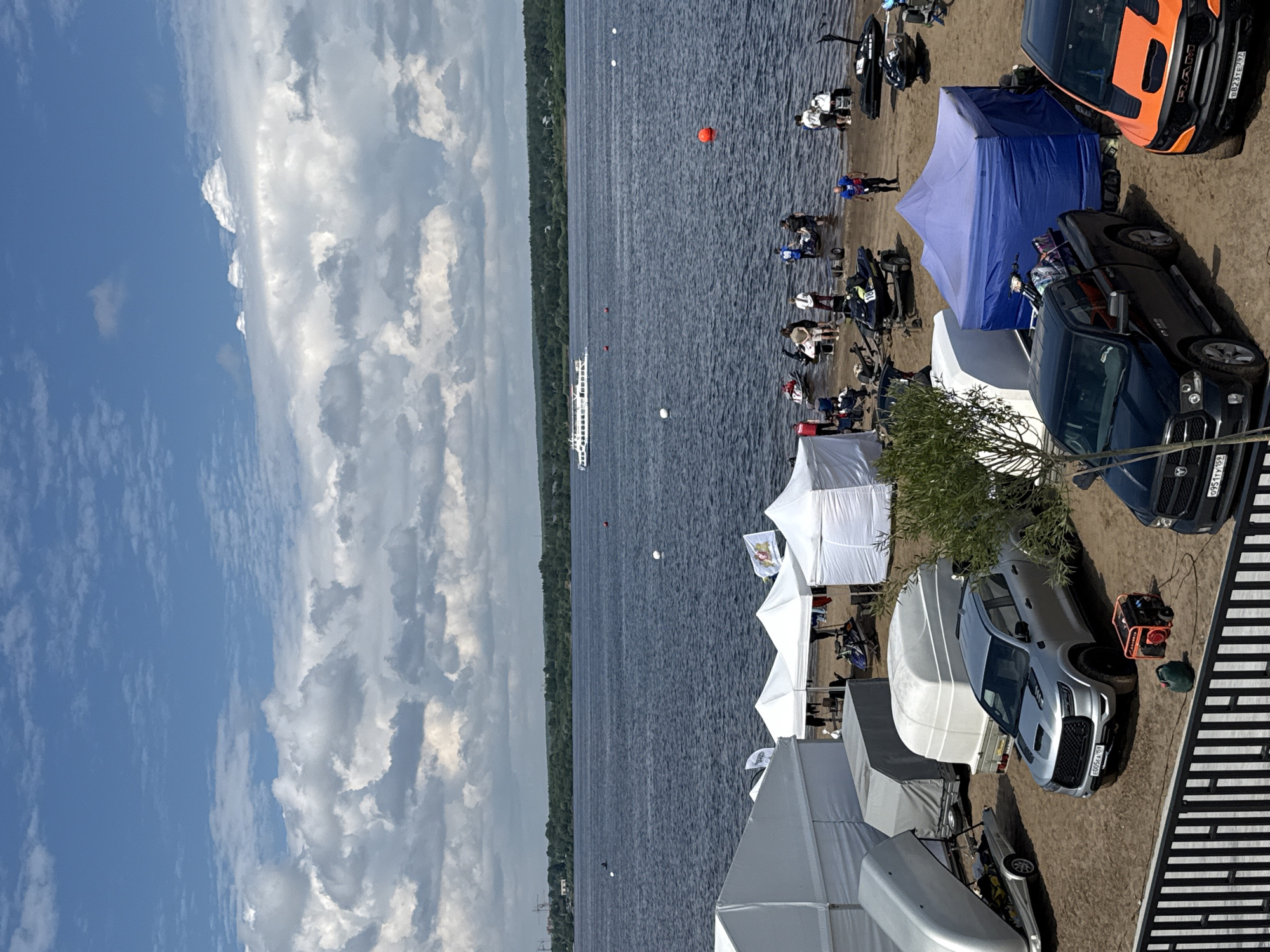 The image depicts a sunny day on the Kama Riverfront in the city of Perm, Russia, likely at the location of a water sports event. In the foreground, several white tents and a large blue tent are set up on a sandy beach. Various vehicles, including trailers with boats or watercraft, a silver pickup truck, a black pickup truck, and an orange pickup truck, are parked on the sand. A portable generator and a gas can are visible near the silver truck.
The middle ground shows a group of people gathered on the sandy bank of the river, some standing around jet skis and other watercraft. Several jet skis are on trailers or directly on the sand. Buoys are scattered in the river, marking a course or area for water activities. In the background, the wide Kama River stretches out, with a large white passenger boat sailing in the distance. The opposite bank of the river is lined with a dense green forest, and a hazy skyline with industrial structures and cranes is visible on the far horizon. The sky is partly cloudy with large, dramatic white cumulus clouds against a bright blue expanse.