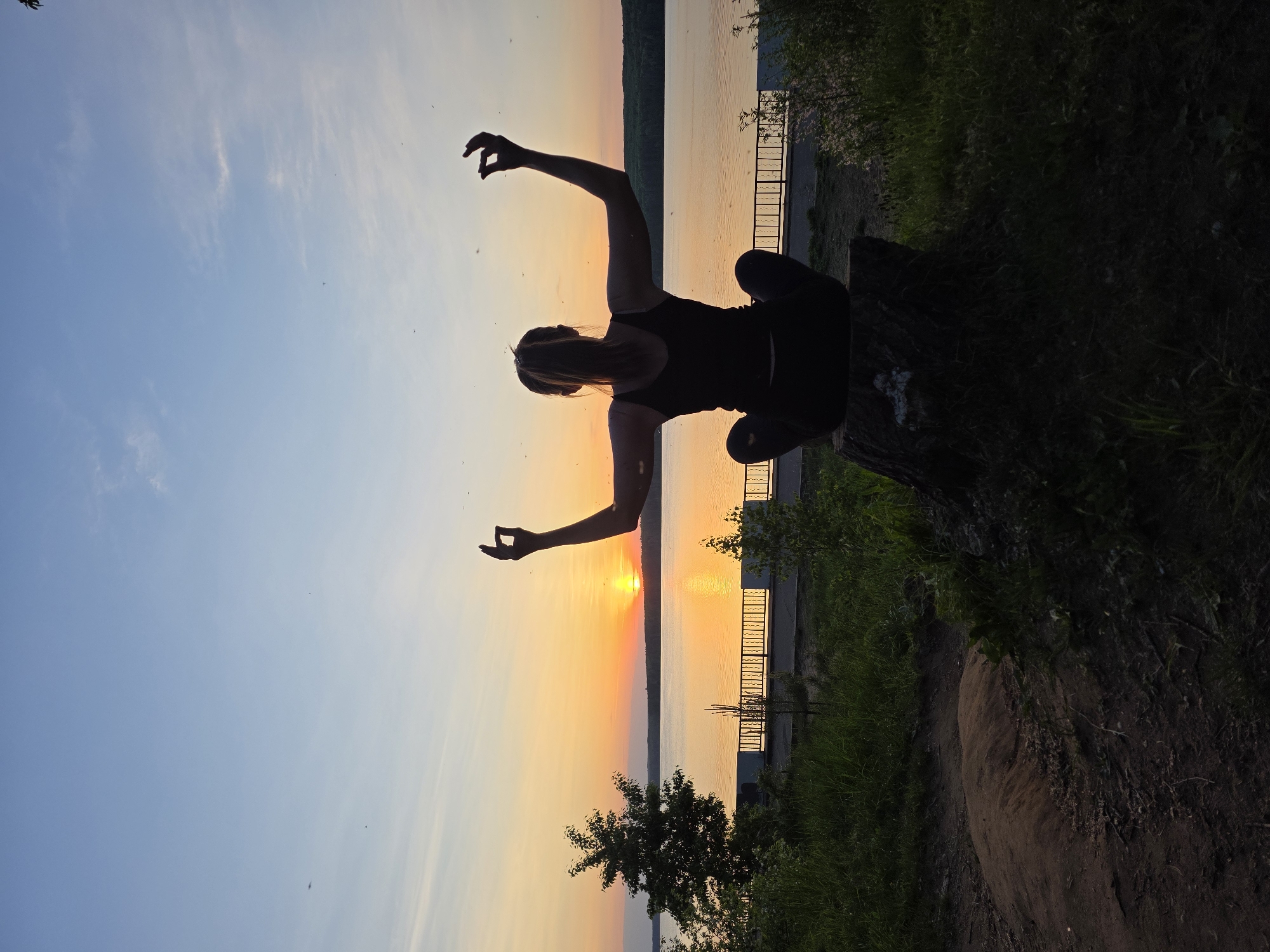 A person in silhouette sits cross-legged on a tree stump at sunset. The person is likely female with blonde hair and is wearing dark clothing. Her arms are raised with hands in a gesture that could be a mudra or an expressive pose. The background consists of a body of water reflecting the sunset. The opposite shore is visible in the distance. A metal fence can be seen near the shore in the middle ground. Vegetation is present on the right side of the frame, further suggesting a natural, outdoor environment. The scene is possibly located in the city of Chaikovsky, Russia.