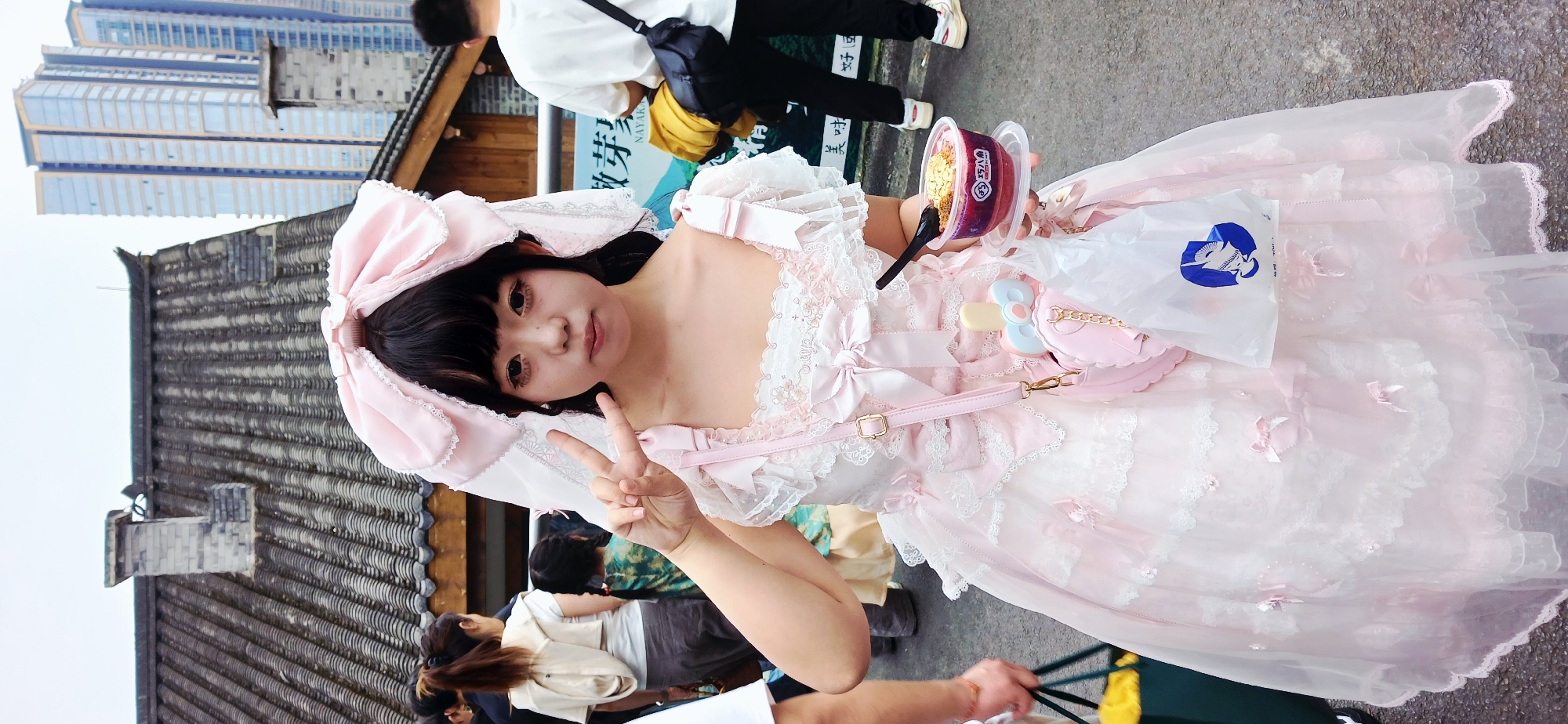 A young person dressed in a pink and white Lolita-style dress and a matching bonnet is seen outdoors, holding a cup of a dessert-like food item and a plastic bag with a blue logo. The individual is making a peace sign with their hand. Other individuals are visible in the background, along with a building with traditional Chinese-style rooftops and a modern high-rise in the distance. The scene appears to be on a city street within the Yuzhong District of China.