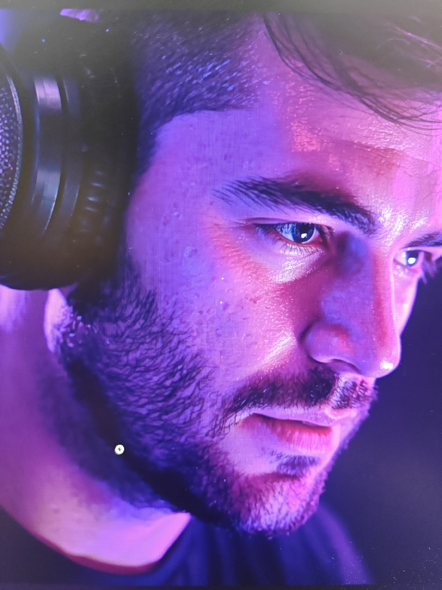 The image is a close-up, low-angle shot of a person wearing headphones and facing to the right. The subject appears to be a male with a beard and short hair, with sweat visible on his face. The lighting is predominantly purple and pink, creating a moody and intense atmosphere. The headphones are black and cover the subject's left ear. The background is dark and out of focus. There are no identifiable landmarks or contextual clues that would indicate a specific location within the city of Yekaterinburg, Russia.