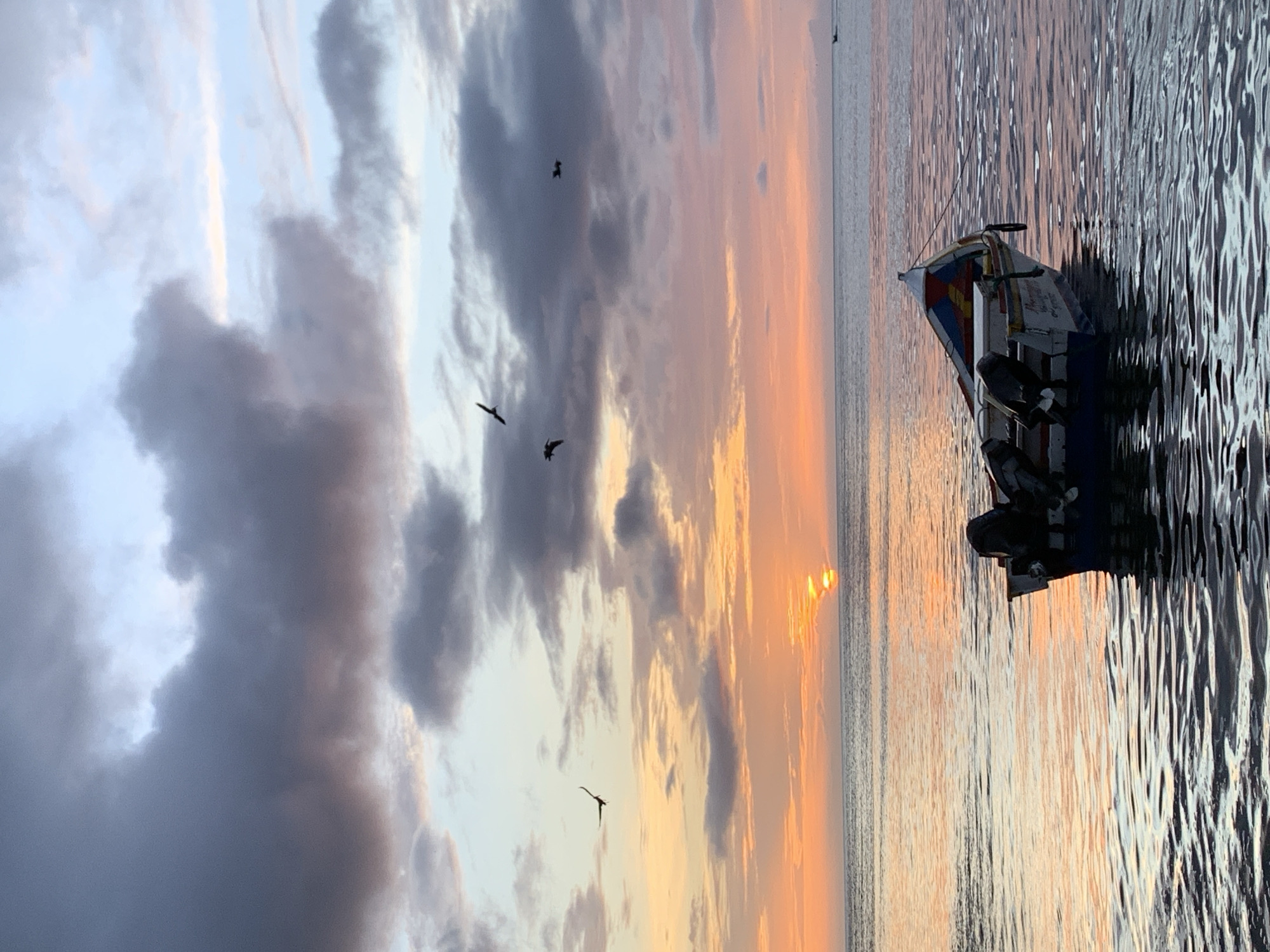 A wide-angle, eye-level photograph captures a tranquil scene at sea in Juan Griego, Venezuela, during what appears to be either sunrise or sunset. The sky is a canvas of soft, blended colors, transitioning from a pale blue at the top to vibrant oranges and pinks closer to the horizon. Dark, textured clouds are scattered across the sky, with lighter patches suggesting sunlight breaking through.
In the middle ground, the calm sea reflects the colors of the sky, with gentle ripples creating a shimmering effect. A small, motorized boat, painted white with blue, red, and yellow accents, floats serenely on the water. The boat is equipped with two outboard motors and is tethered. A faint inscription is visible on the side of the boat.
Several birds are depicted in silhouette against the sky, with four birds clearly visible in flight. The sun is a bright, diffused orb just above the horizon, casting a golden glow over the water. In the far distance, a faint landmass or large wave is visible on the horizon. The overall impression is one of peace and natural beauty.