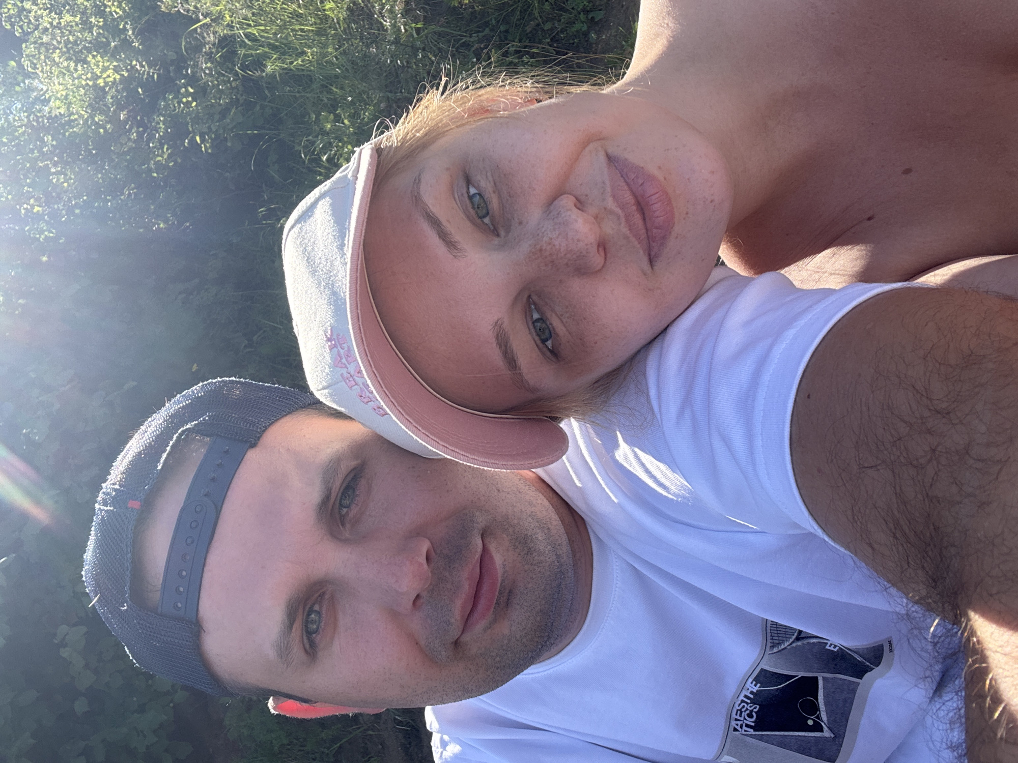 A selfie taken outdoors shows a man and a woman close together. The man, wearing a dark blue baseball cap and a white t-shirt with a graphic on the left side, is on the left of the frame. He has light skin, light blue eyes, and stubble. The woman, on the right, is wearing a light pink baseball cap and a white t-shirt. She has fair skin with freckles, light brown hair, and light blue eyes. She is leaning her head on the man's shoulder. Both individuals appear to be smiling subtly. The background consists of green foliage and grass, suggesting a natural or park-like setting. The sun is shining brightly, creating lens flare in the upper right portion of the image. The lighting and greenery indicate a daytime outdoor setting, potentially during warmer months. The location is identified as Yaroslavl, Russia.