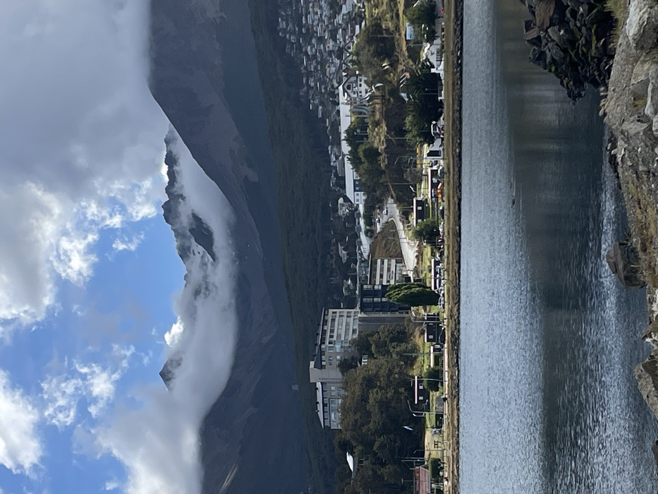Mountains loom large under a partly cloudy sky. One peak is shrouded in mist. The mountains recede into a densely populated town along a narrow shore. A body of water extends toward a rocky edge in the foreground. Buildings line the shoreline, interspersed with trees and streets. The scene is dominated by natural elements, with the town nestling between the mountains and the water.