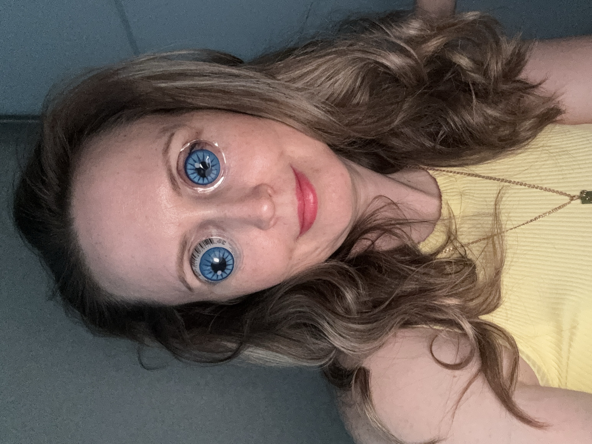 The image shows a woman with brown hair and blue, doll-like eyes that include a barcode around the pupil, wearing a yellow shirt and a gold necklace. There is no information indicating this image was taken in Chaikovsky, Russia.