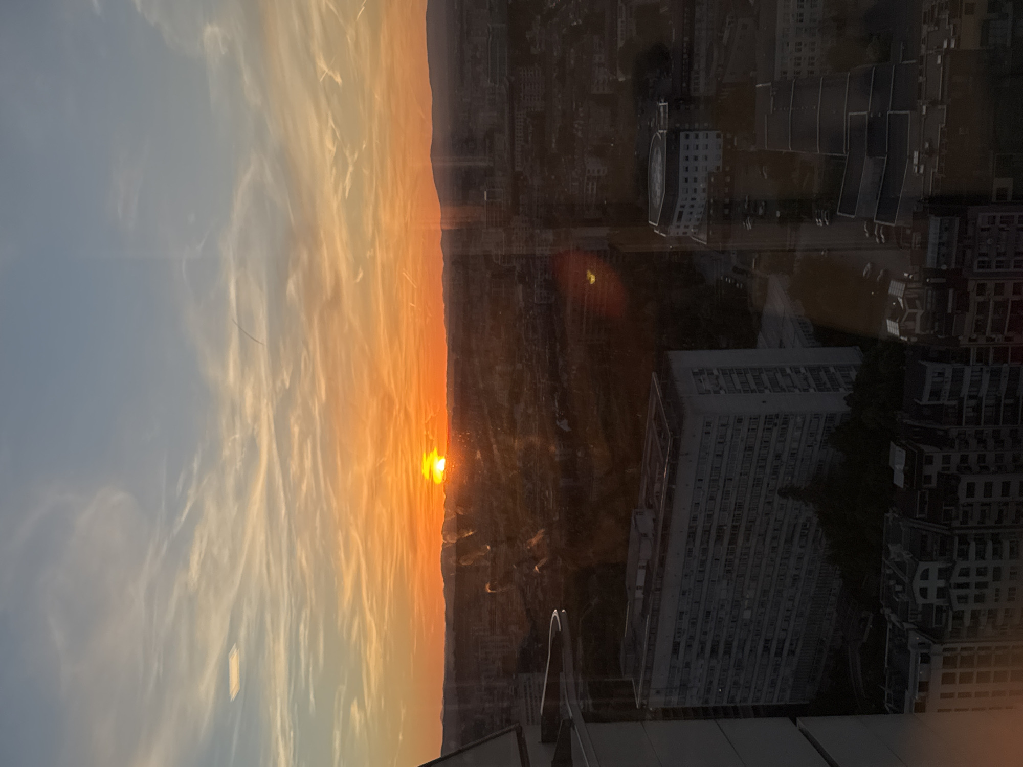 The image depicts a sunset over Sofia, Bulgaria, as viewed from a high vantage point. The sun is setting over a ridge of hills in the distance, casting an orange glow over the sky. In the foreground, reflections of buildings are visible in a window or pane of glass. The image showcases a blend of natural beauty and urban elements.