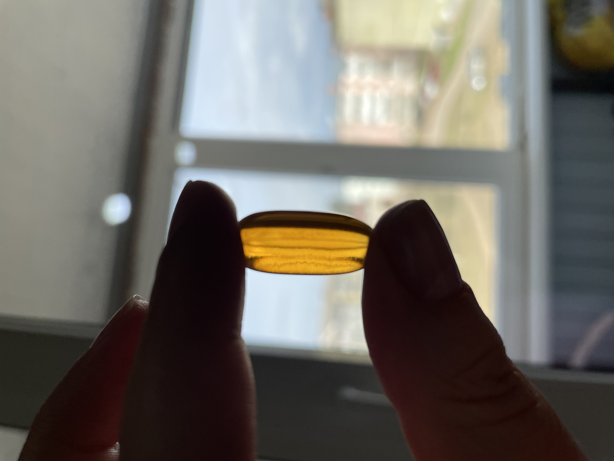 A person holds a translucent, yellow-orange, oblong-shaped softgel capsule between their thumb and index finger. The capsule is held up against a window, which is slightly out of focus, revealing an exterior view of a building. The building appears to have multiple stories and is white or light-colored with a red roof. It cannot be seen, that the location is Noviy, Russia.