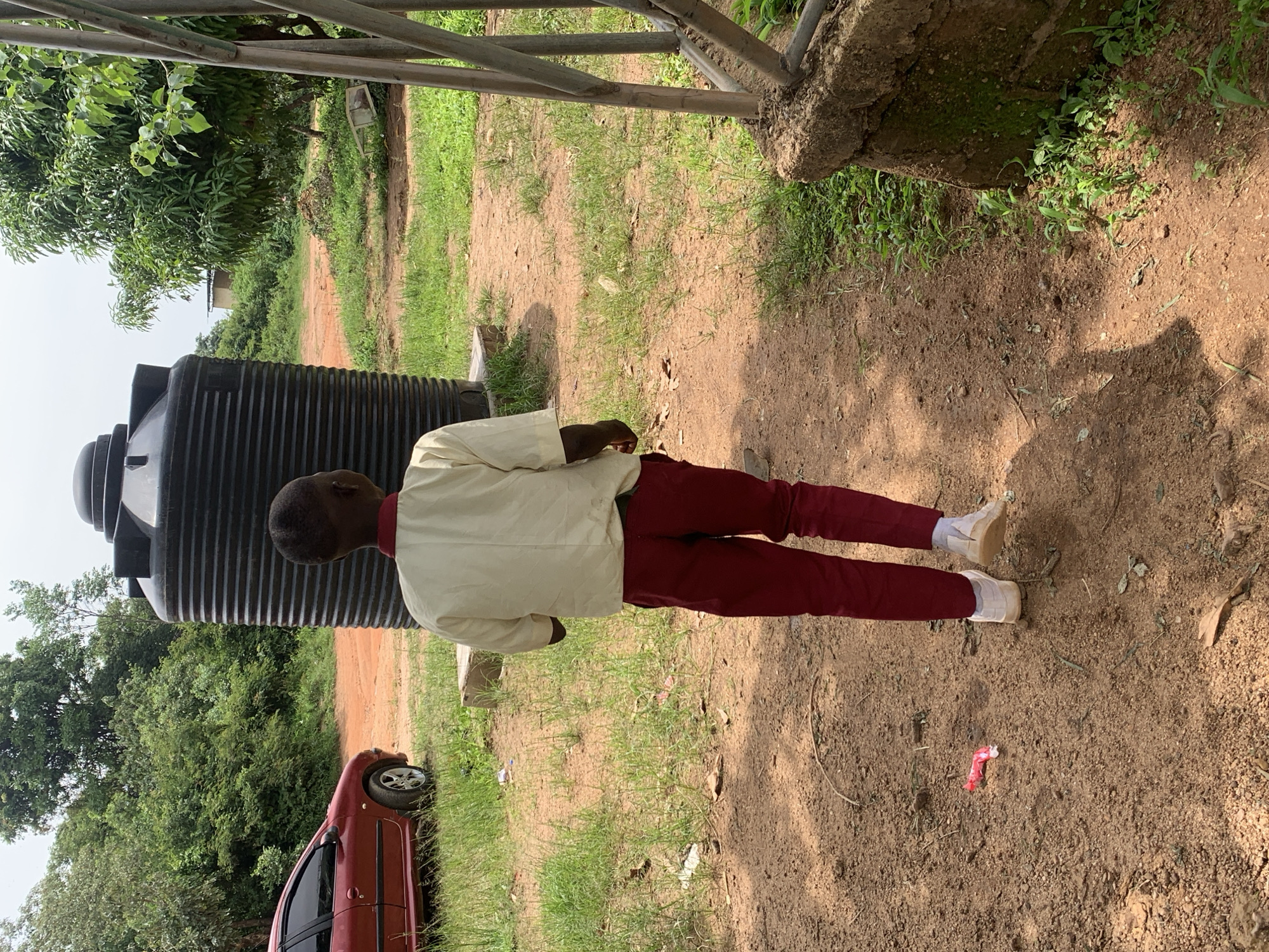 A young boy, viewed from behind, stands on a dirt path in what appears to be an outdoor area in Jos, Nigeria. He is wearing a light-colored short-sleeved shirt and dark red pants, with white shoes. To his left, a large, dark-colored water tank is visible, a common sight in Nigerian communities for water storage. Behind the tank and to the left, a red car is parked. The ground is a mixture of dirt and sparse green grass. In the background, there are trees and some vegetation, and a simple building can be seen further in the distance. The sky is overcast. The scene suggests a potentially rural or semi-urban setting, possibly associated with a school or community center due to the water tank and the child's attire.
