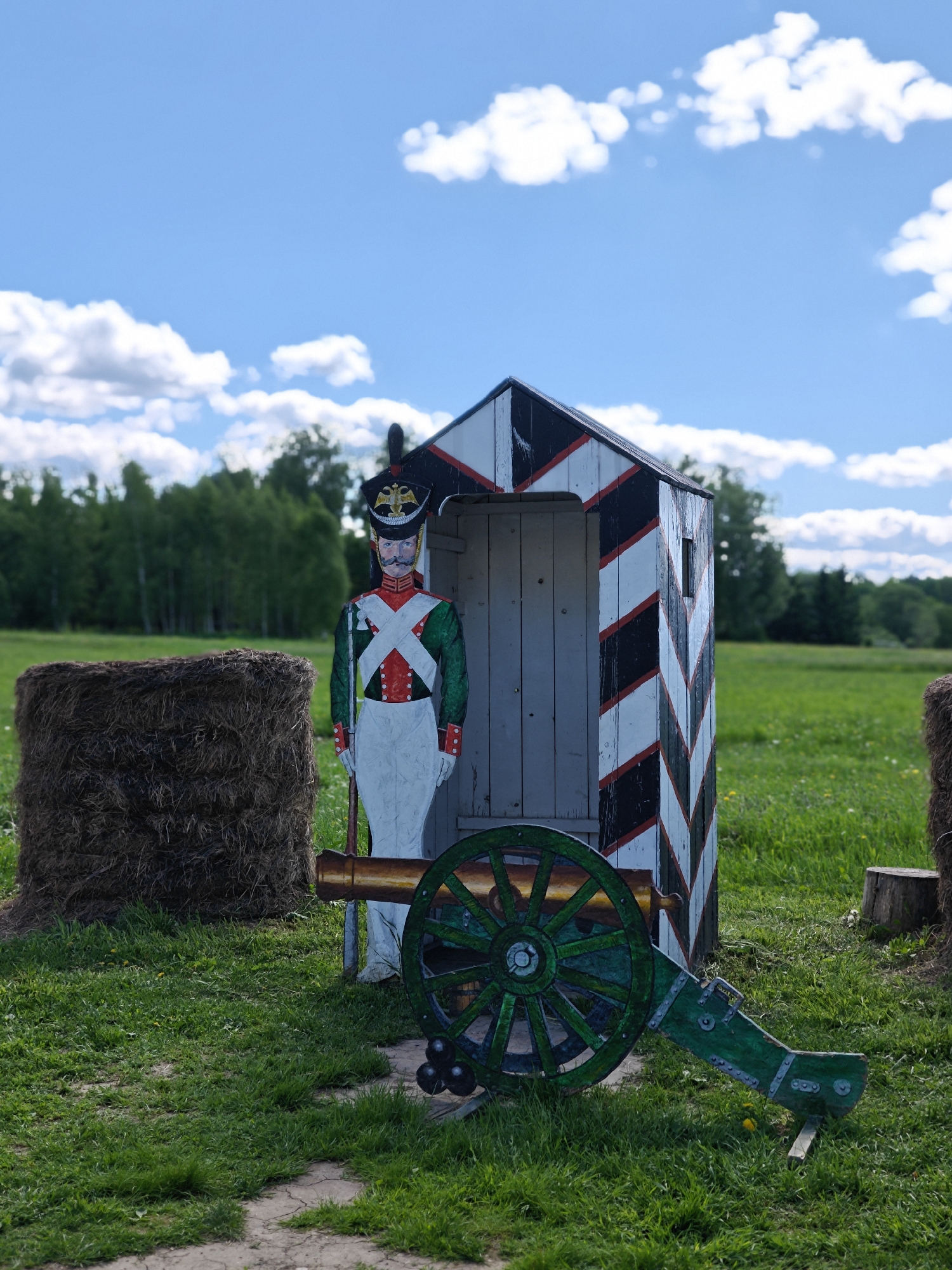 The image depicts a scene at the Borodino battlefield, near Moscow, Russia. A wooden cut-out of a Napoleonic soldier stands guard next to a sentry box marked with black and white chevron patterns. A decorative green cannon with cannonballs sits in front of the soldier and box. A hay bale is positioned to the left of the box, and a field with trees is visible in the background under a partly cloudy blue sky.
