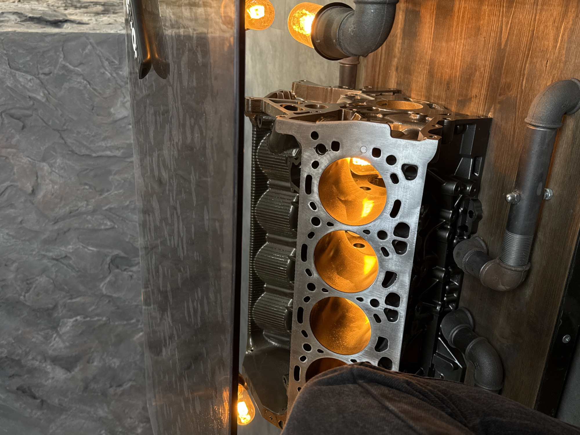 The image shows a close-up of an engine block used as a decorative element. The block is made of metal, with visible cylinder openings illuminated with a warm orange light source. The block is mounted on a wooden panel with black pipes running alongside. The lighting fixture includes exposed bulbs. The image includes a partial view of a person's clothing near the bottom of the image. The location and the city of Кондратово, Russia are not identifiable from the image.