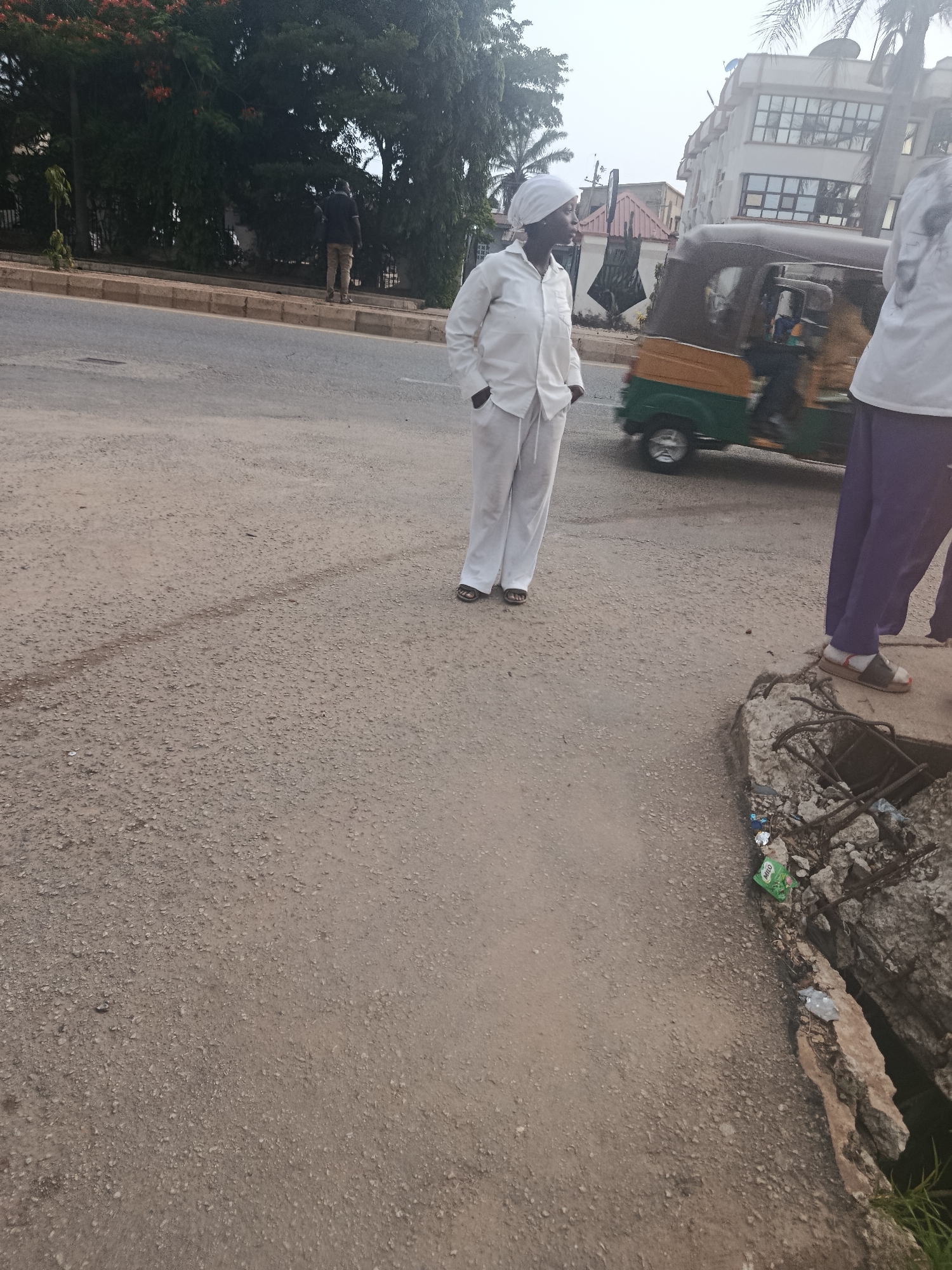 A person wearing a white headwrap and a white outfit stands on a dusty road in New Stadium, Nigeria. The person is facing left, with hands in their pockets. Behind them, a three-wheeled vehicle, likely a tuk-tuk, moves along the road. The background includes buildings and trees. To the right, another person wearing purple pants and a white top is partially visible, standing near a broken concrete structure. The ground is unpaved and appears to be under construction or in disrepair, with debris scattered around. The lighting suggests it is daytime.