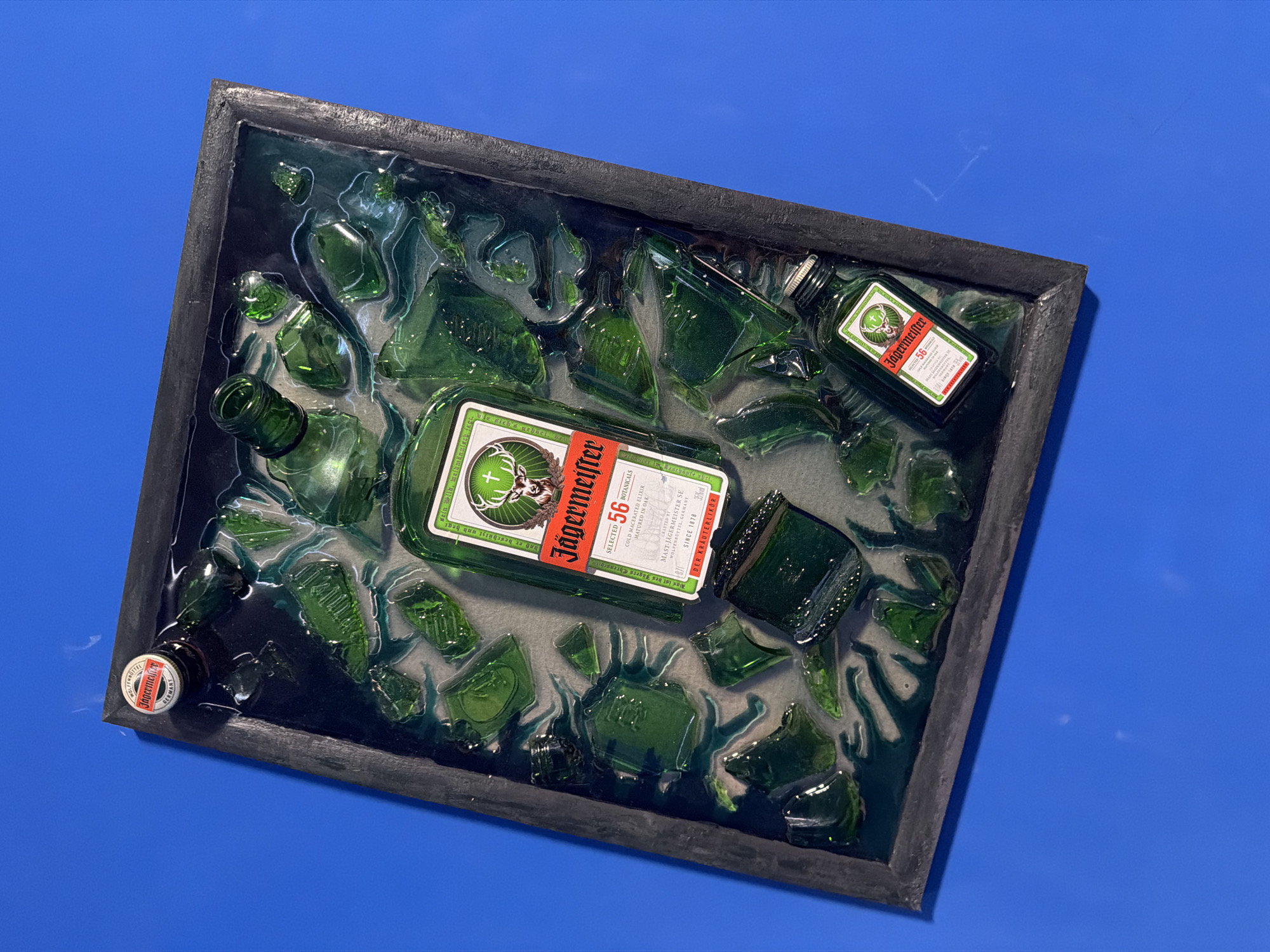 A top-down, slightly angled image captures a framed artwork or display, likely located in Chaikovsky, Russia. The central subjects are a large Jägermeister bottle, a miniature Jägermeister bottle, and numerous fragments of broken green glass, all encased within a dark, translucent, greenish-blue resin. This resin fills a rectangular frame with a dark, textured finish.
The full-sized Jägermeister bottle lies diagonally across the middle of the frame, with its iconic red and white label prominently displayed. A smaller Jägermeister bottle is visible in the upper right corner, also showcasing its label. In the lower-left corner, a Jägermeister bottle cap is partially visible. The broken glass pieces are distributed throughout the resin, suggesting they are submerged or permanently set within the solidified material, which exhibits a subtle wavy texture.
The framed piece is presented against a plain, vivid blue background. The lighting is uniform, indicating an indoor setting.
Visible text on the labels of the Jägermeister bottles includes the brand name "Jägermeister," the number "56" (denoting botanicals), "SELECTED BOTANICALS," "COLD MACERATED ELIXIR," "CRAFTED BY MAST-JÄGERMEISTER SE WOLFENBÜTTEL, GERMANY," "SINCE 1878," "35% VOL," and "DER KRÄUTERLIKÖR." A German hunting motto, "Was ist des Jägers Ehrenschild, daß er beschützt und hegt sein Wild, weidmännisch jagt," is also legible on the main label. The cap on the smaller bottle also states "WOLFENBÜTTEL GERMANY."