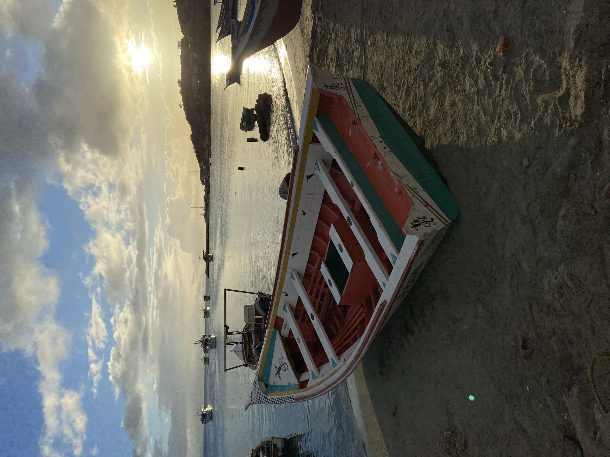 In Juan Griego, Venezuela, a colorful wooden fishing boat rests on a sandy beach near the water's edge. The boat, painted in red, white, green, and yellow, faces towards the sea. Other boats, including larger vessels, are anchored in the calm water. The sun is setting, casting a warm glow on the scene and reflecting on the water, with a hillside dotted with buildings and vegetation visible in the background. The sky is a mix of blue with scattered clouds. The sand is wet near the water and shows some textures and debris, including a small red object and a tiny green light. The overall impression is a peaceful evening by the coast.