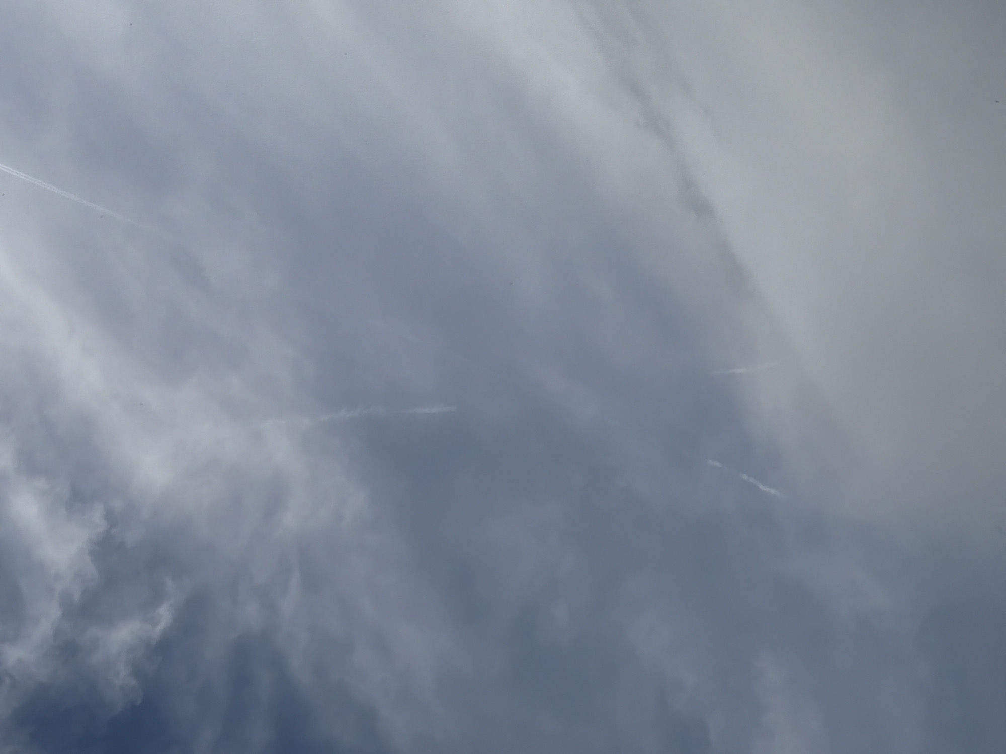 The image shows a cloudy sky. Three contrails are visible, stretched across the sky, created by passing aircraft. The sky is a mix of grey and white clouds with patches of blue. No people or objects on the ground are visible. There is no way to verify the location of the picture.
