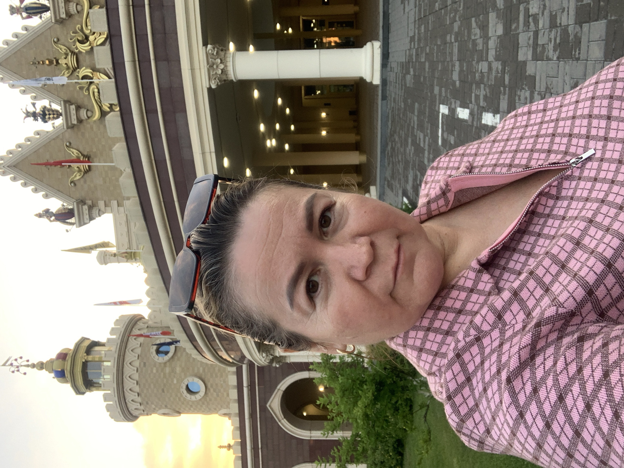 The image shows a woman taking a selfie in front of the "Pyramid" shopping and entertainment complex in Kazan, Russia. The woman is wearing a pink and brown checkered jacket and sunglasses are perched on her head. The building behind her features a distinctive architectural style with towers, flags, and ornate decorations. The sky in the background suggests it is either sunrise or sunset, with a warm golden glow. The foreground shows a paved area with white markings, likely a walkway or parking area, and some greenery to the left.