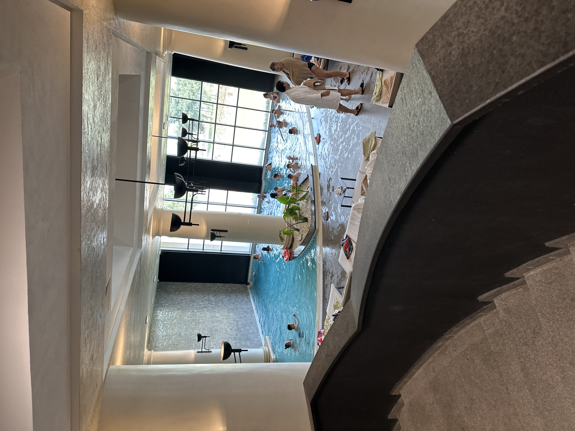 The image captures a high-angle view of an indoor swimming pool area, possibly a spa or wellness center, in Kazanlak, Bulgaria. The architecture is modern with tall windows and pillars. Several people are in the pool, while others are lounging on chairs or walking along the poolside. Some individuals are wearing robes. Tables and plants decorate the area. The viewpoint appears to be from a higher level, possibly a staircase, with the steps visible in the foreground.