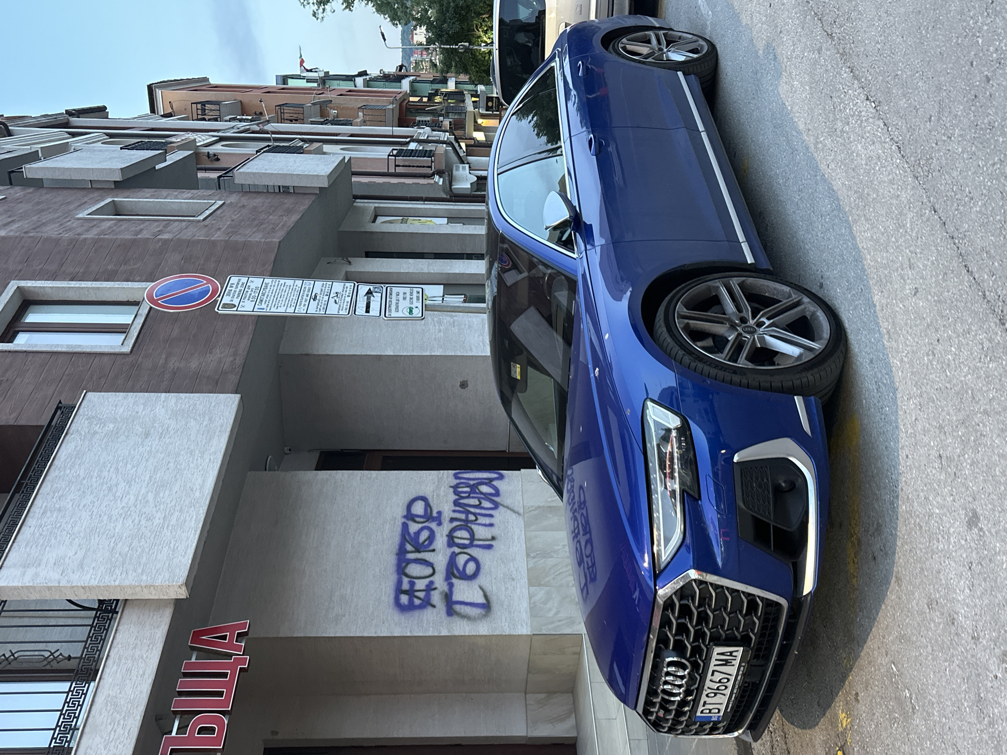 The image shows a blue Audi A8 parked on a street in Veliko Tarnovo, Bulgaria. The car is in the foreground, with a building behind it. The building has a sign with red Cyrillic letters that reads "ЛЪЩА". There is graffiti on the wall of the building that also appears to be in Cyrillic. A parking sign is visible on the building, and several other buildings with balconies and colorful facades are visible further down the street. The sky is overcast. The license plate of the car is visible and reads "BT 9667 MA".