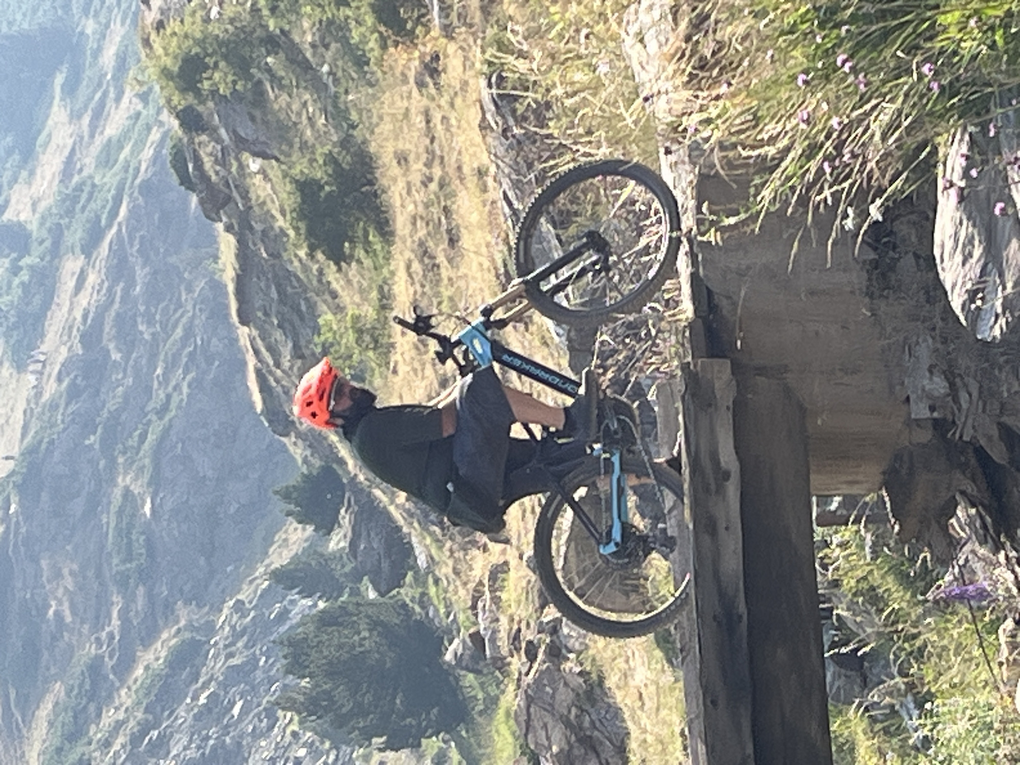 A man wearing a black t-shirt, black shorts, and an orange helmet is riding a mountain bike over a wooden bridge. The bike is black and blue with yellow accents. The man is looking to his right. The bridge is old and made of rough-hewn wood. The background is a rocky, mountainous landscape with dry grass and some green bushes. In the far background, there are more mountains and trees. The sky is clear and blue. This scene appears to be located in the Pyrenees mountains, a region that includes Benasque, Spain.