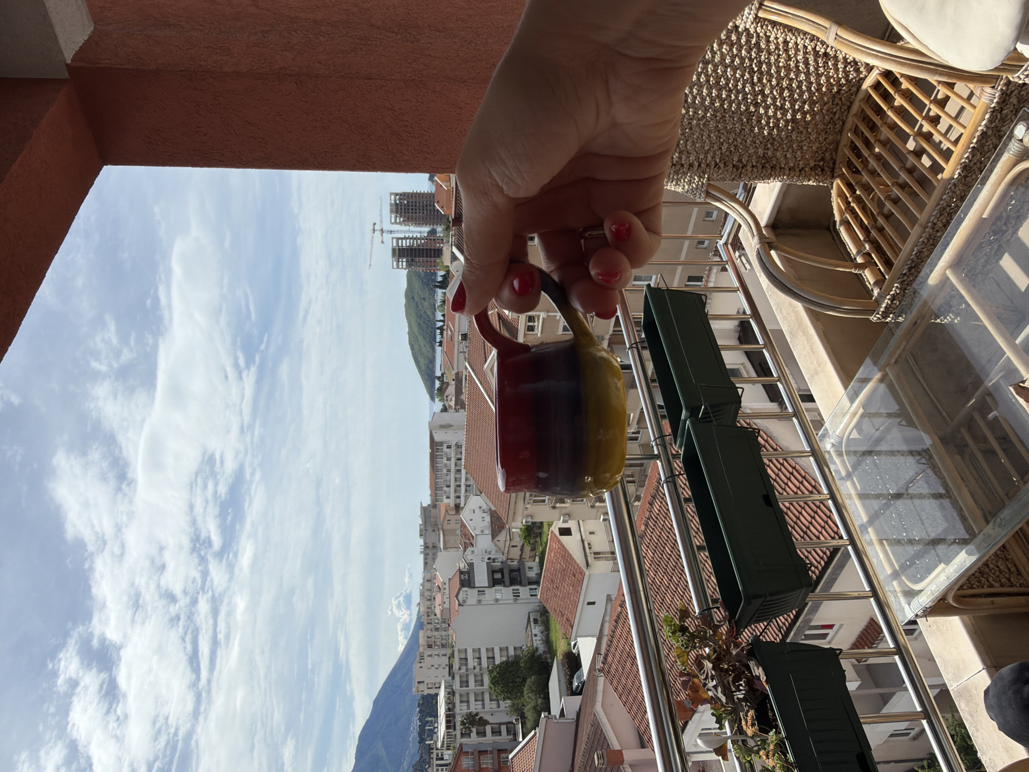 The image captures a vibrant, sunny daytime view from a balcony overlooking a bustling coastal city, identified as Budva, Montenegro. In the foreground, a person's hand, adorned with red nail polish, holds a multi-colored mug (red, dark purple, and yellow bands) above the balcony railing.
The balcony itself features a modern metal railing with horizontal bars, from which several dark green rectangular planter boxes are suspended. To the right, a woven rattan chair with a light cushion and a glass-topped table are visible, suggesting an outdoor seating area. The balcony wall and floor are textured and appear to be a warm terracotta or reddish-brown color.
Beyond the balcony, a dense cityscape unfolds with numerous buildings, predominantly white or light-colored, featuring distinctive red or terracotta tiled roofs. This architecture is characteristic of the Mediterranean region. In the background, rolling green hills and mountains rise majestically, adding to the scenic beauty. Several tall buildings under construction, marked by visible cranes, indicate ongoing development in the area. The sky above is a bright, clear blue with scattered white clouds, suggesting pleasant weather. No visible text is present in the image.