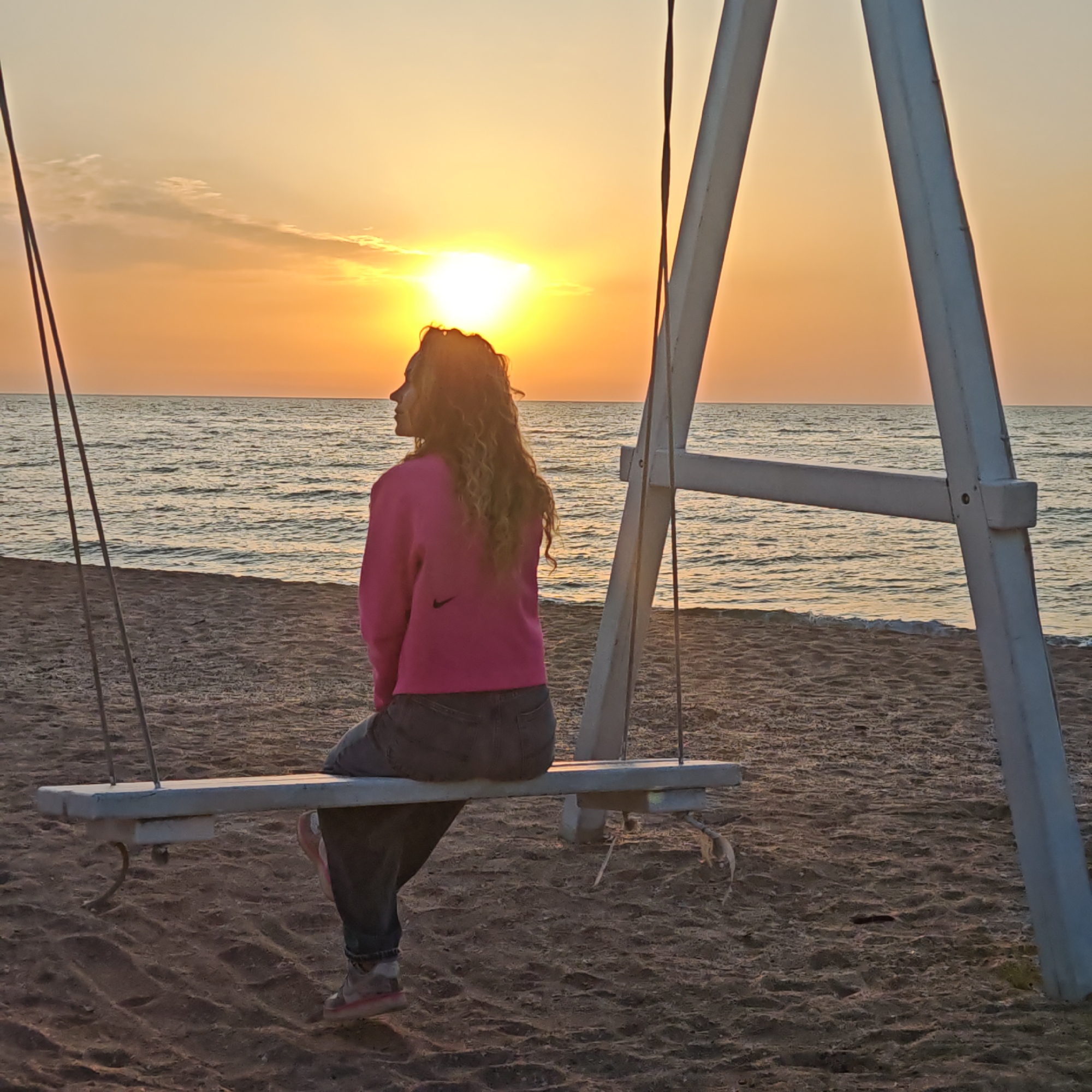 A woman sits on a swing on a beach at sunset in Мысовское сельское поселение, Ukraine. The woman faces the ocean. The swing has a white frame. The beach is sandy. The sun is setting over the water. The woman wears a pink sweatshirt and dark jeans.
