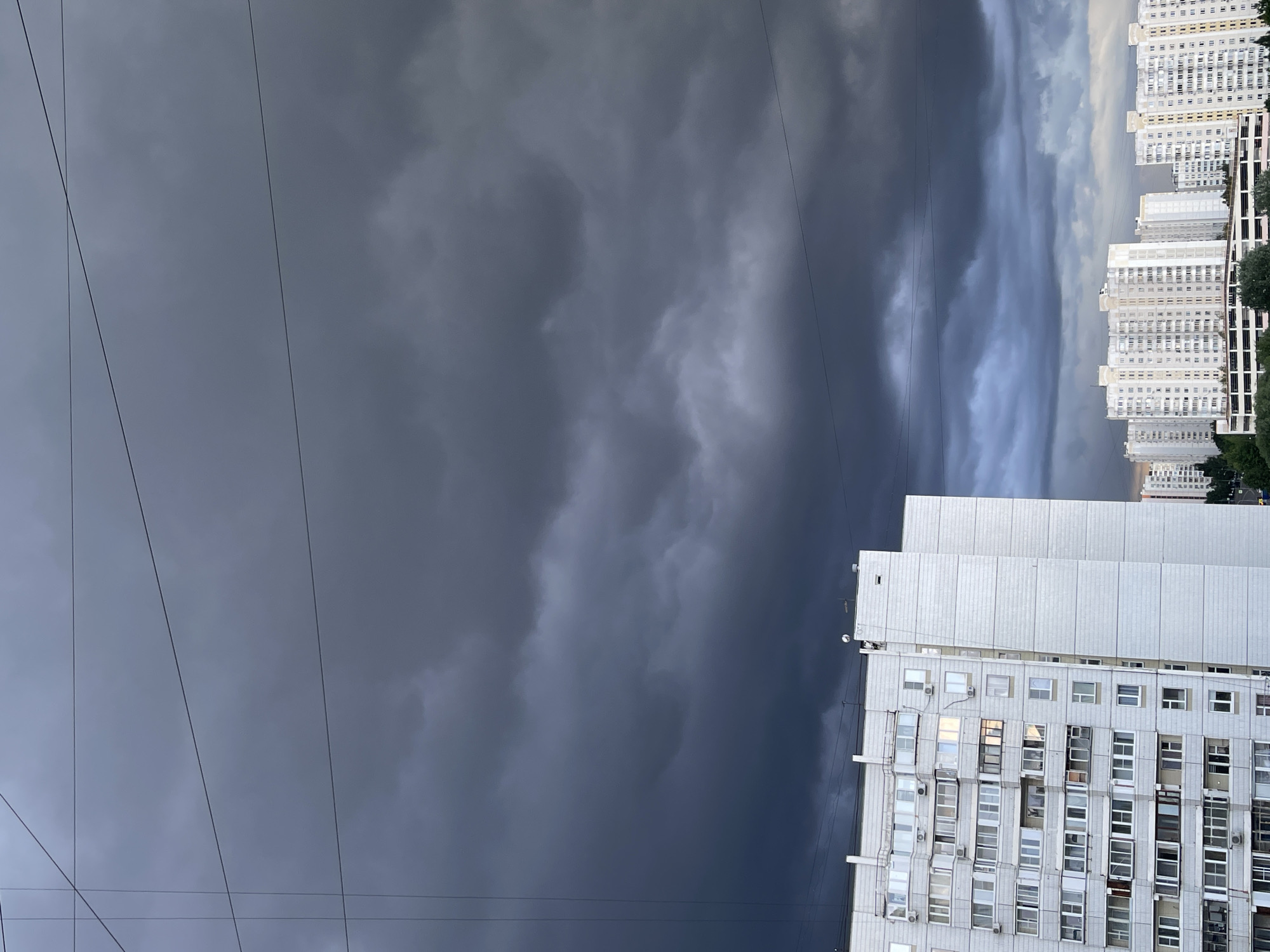 The image depicts a cloudy sky above a city, likely in Moscow, Russia, as inferred from the architectural style of the buildings. The clouds range from dark gray to lighter shades, suggesting an approaching storm. The buildings are multi-story residential blocks typical of Soviet-era construction. There are power lines in the foreground. No people are visible.