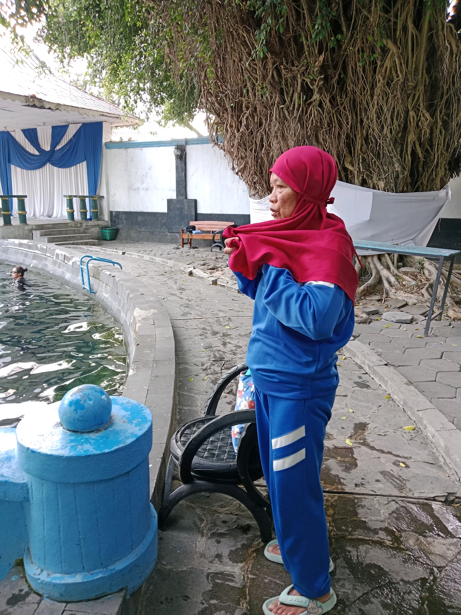An adult woman wearing a red hijab and a blue tracksuit stands on a paved area next to a swimming pool. The woman is facing to her right and adjusting her tracksuit top. To the left of the woman, a swimming pool filled with water is visible, with a person swimming in it. A blue railing runs along the edge of the pool. In the background, a white wall is decorated with blue and white drapes, and large tree roots are visible. The scene appears to be outdoors in a recreational area. The location is identified as Jentekan, Indonesia.