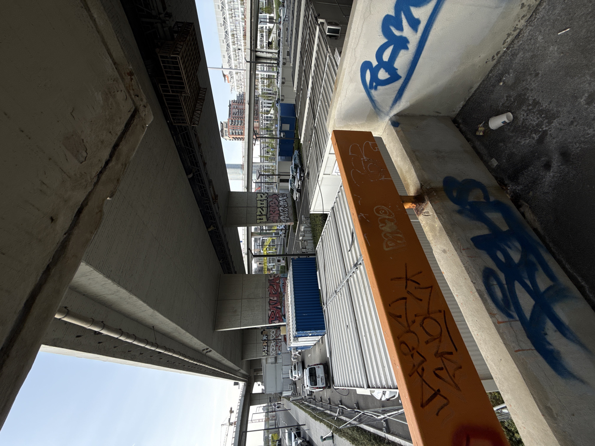 The image, captured from an elevated perspective and rotated, depicts an urban scene beneath a large concrete overhead structure, likely an elevated road or railway, in Belgrade, Serbia. The view looks down onto a street-level area.
On the left, the underside of a massive light-colored concrete beam dominates, showing textured surfaces and utility conduits with small lights or sensors. On the right, a thick concrete support column features bold blue spray paint graffiti. In front of this column, an orange-painted metal beam or barrier is also covered in graffiti, where the word "ROCK" is clearly visible, among other illegible tags. Litter, including a white paper cup and cigarette butts, is scattered on the dark ground next to the column.
Below the overhead structure, a bustling temporary or industrial zone unfolds. Numerous white shipping containers are arranged along the street, some fitted with air conditioning units, suggesting use as temporary offices or facilities. A prominent blue shipping container stands out amidst the white ones. Several white vans and at least one white car are parked or driving on the asphalt road below. Further along the left, another concrete wall is heavily adorned with various graffiti tags.
In the background, a clear blue sky is visible, indicating bright daytime conditions. Distant multi-story buildings, along with a large white metallic structure (possibly part of a bridge or another overhead construction), frame the urban landscape. A faint construction crane is visible in the far background on the lower left. No people are visible in the scene.