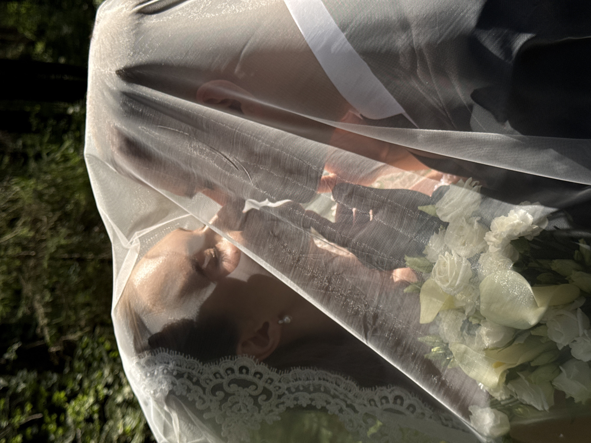 A bridal couple, partially obscured by a veil, are shown in a close-up shot outdoors. The bride, on the left, is wearing a white veil with lace trim and pearl earrings. Her eyes are closed as she leans in to kiss the groom. The groom, on the right, is wearing a dark suit and a white shirt. A bouquet of white flowers, including roses and calla lilies, is visible in the foreground, also partially covered by the veil. The background consists of lush green foliage, suggesting a natural, outdoor setting. The lighting indicates it is daytime, with sunlight filtering through the trees and the veil, creating soft shadows and highlights. The image is taken from a medium distance, focusing on the couple's embrace.