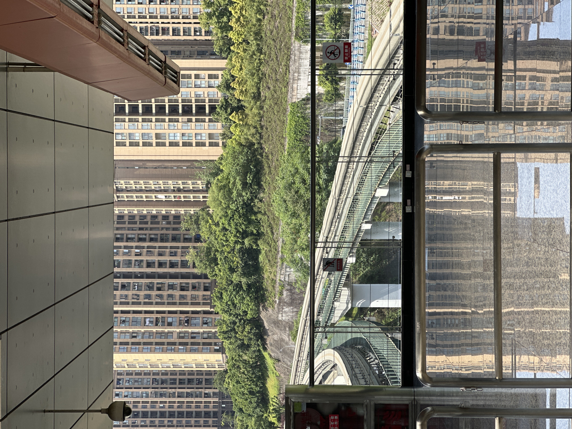 The image depicts an elevated view from a building in the city of Jiansheng, China.
The main subjects are:
1. A tall, beige building with many windows, repeated in a reflection in the right-hand side of the image.
2. Lush greenery and a concrete hillside.
3. An elevated, curved rail track system.
4. A building with a gray tiled exterior, connected to an overhanging brown structure with horizontal pipes.
A red and white sign with a pedestrian crossed out is visible next to the rail track. In the lower right corner is a glimpse of a red object behind glass, possibly a fire extinguisher.