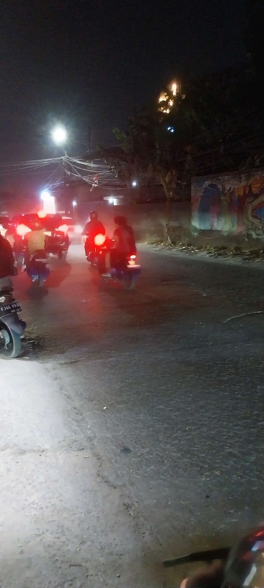 In the city of Bojong Nangka, Indonesia, a nighttime scene unfolds on a street at an intersection. A street lamp illuminates the area with a bright white light, casting long shadows and highlighting a dense network of electrical wires overhead. To the right, a wall is adorned with a colorful mural. The road is wet, reflecting the lights of vehicles. Several motorcycles are present, their red taillights creating streaks of light on the asphalt, suggesting movement. One motorcycle in the foreground has a license plate visible, reading "FJ66 RRB." The overall atmosphere is one of a busy, active street at night.