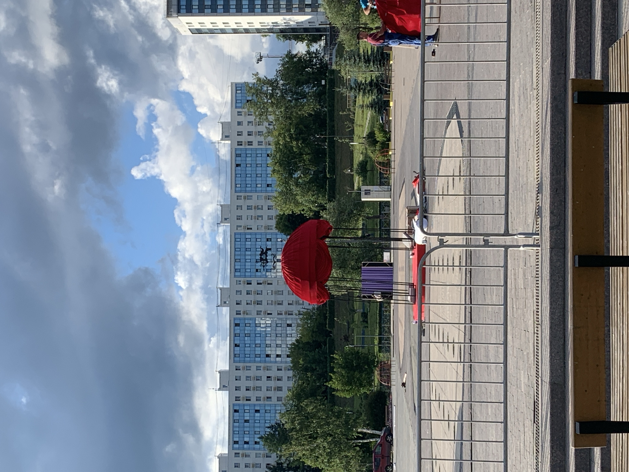 The image depicts an outdoor scene in front of a modern, multi-story building complex, characteristic of a city like Perm, Russia. In the foreground, a metal fence separates the viewer from a paved area. Beyond the fence, a red, umbrella-like structure is the central focus, supported by poles and suspended over a circular red platform. A purple curtain is visible behind this structure.
To the right of the red structure, two individuals are present. One, wearing a blue shirt and jeans, appears to be interacting with a red fabric, possibly setting up or taking down equipment. Another individual, in a blue top and dark pants, is also engaged in activity near the red fabric. Several other objects are scattered on the paved area, including what appears to be a red car in the background to the left.
The environment includes green trees and a grassy slope behind the paved area, leading up to the residential buildings. The sky is partly cloudy, with a mix of dark and light clouds. A tall, modern building stands to the far right, also featuring a facade of windows. The overall scene suggests an outdoor event or performance area being prepared or managed. The presence of the large apartment buildings and the general urban landscape strongly indicates a location within a city, and given the context, likely Perm, Russia.