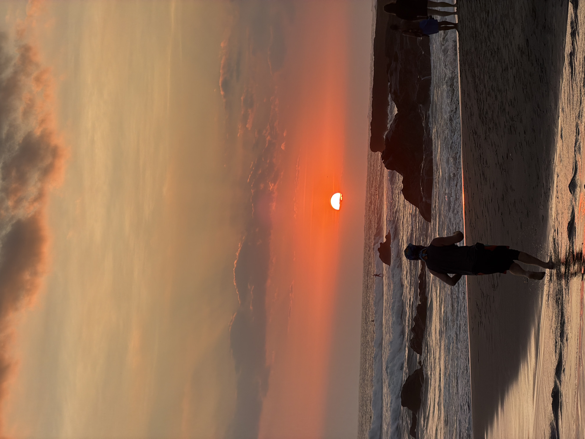 The image captures a sunset at Echo Beach, a popular surfing spot in North Kuta, Indonesia. The sky is a vibrant canvas of oranges and reds, with the sun a glowing orb partially submerged below the horizon. Clouds are scattered across the sky, tinged with the warm hues of sunset.
On the beach, a lone figure is seen running towards the water, silhouetted against the colorful sky. Their stride is energetic, suggesting a determined pace. Further down the beach, two other figures stand near the water's edge, their backs to the camera, observing the sunset. The waves are breaking on the shore, creating a foamy white contrast against the dark sand. Rocks jut out from the water and the shoreline, adding texture and depth to the scene. The wet sand near the water reflects the warm light of the sunset, creating a shimmering effect. In the distance, several surfers can be seen riding the waves, enjoying the last light of the day. The overall atmosphere is one of tranquility and natural beauty.