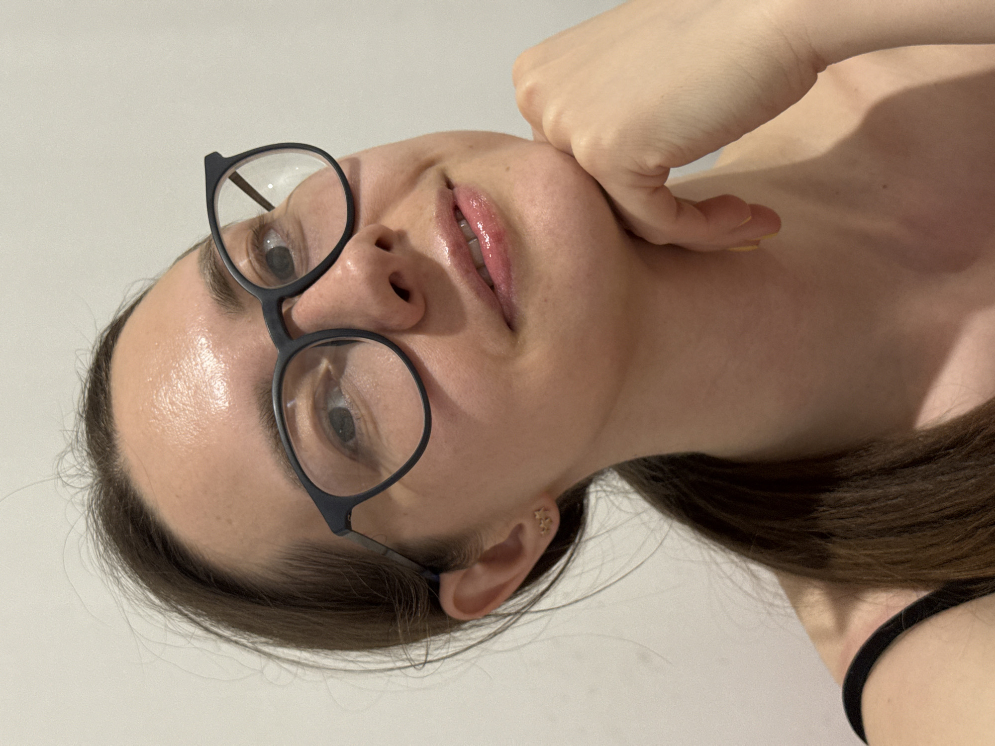 A woman with brown hair and fair skin is shown from a low angle, facing the camera. She is wearing black framed glasses and has gloss on her lips. Her right hand is touching her cheek. She appears to be wearing a black top. A small star earring is visible in her right ear. The background is a plain white wall. The image location is not discernible from the provided information and image.