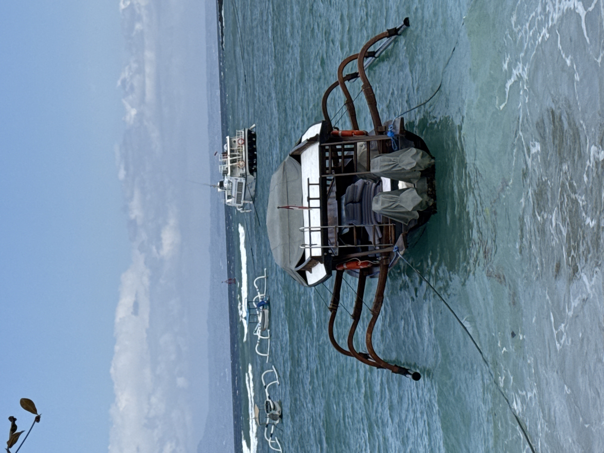 The image shows a body of water, likely the sea, near Manggis, Indonesia. In the foreground, a traditional outrigger boat, covered with a gray tarp, is tied to the shore with ropes. Its distinctive wooden outriggers extend from the sides. In the middle ground, two smaller outrigger boats are visible, also with white outriggers. Further in the background, a larger white passenger ferry is present, with several decks and windows. The water is a vibrant turquoise, with whitecaps indicating waves. The sky is a pale blue with scattered white clouds. The shoreline is out of focus, suggesting the image was taken from the water or close to it.
