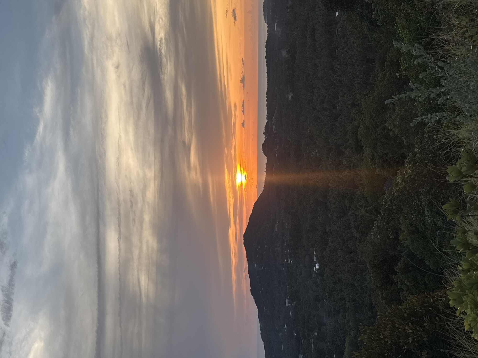 A sunset is visible behind a mountain range in Baan Nai Suan, Thailand. The sky is a mix of light gray, white, and orange clouds, transitioning from lighter shades on the left to more vivid orange hues near the setting sun. The sun is positioned on the horizon, appearing to sit directly atop the mountain peak. A light beam extends downward from the sun towards the trees. The mountain range is densely covered with trees and vegetation.