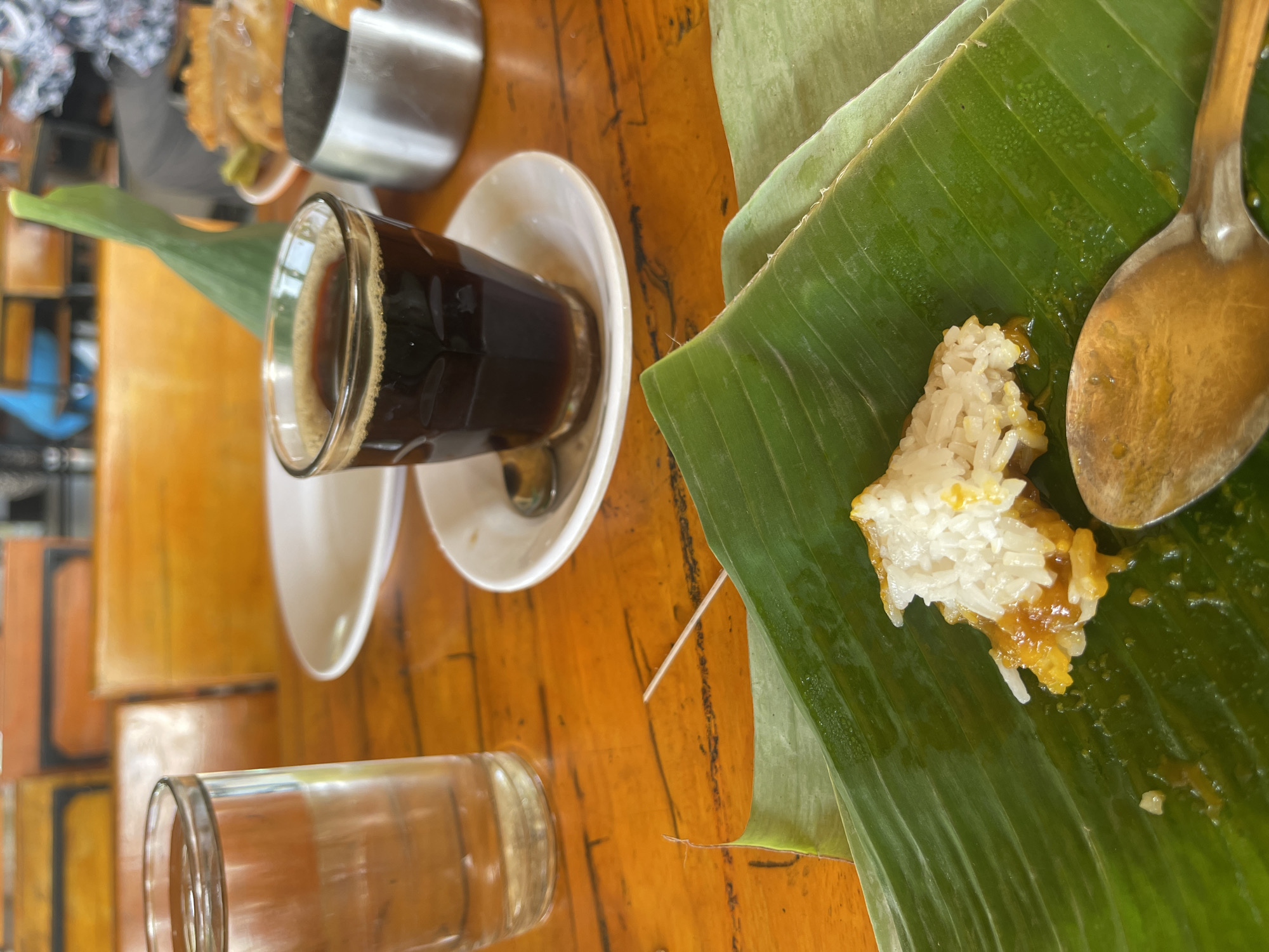 A close-up shot captures a breakfast setting on a wooden table. A clear glass of water sits to the left, partially obscuring some out-of-focus orange seating in the background, possibly within the city of Julikeude Dua, Indonesia. In the center, a small cup of dark coffee rests on a saucer, accompanied by a spoon. To the right, a metal ashtray is visible. The foreground is dominated by a serving of rice on a banana leaf. A small portion of the rice, glistening with a yellow sauce, is positioned on the leaf, with a spoon resting nearby. The scene suggests a casual meal, likely at a local eatery.
